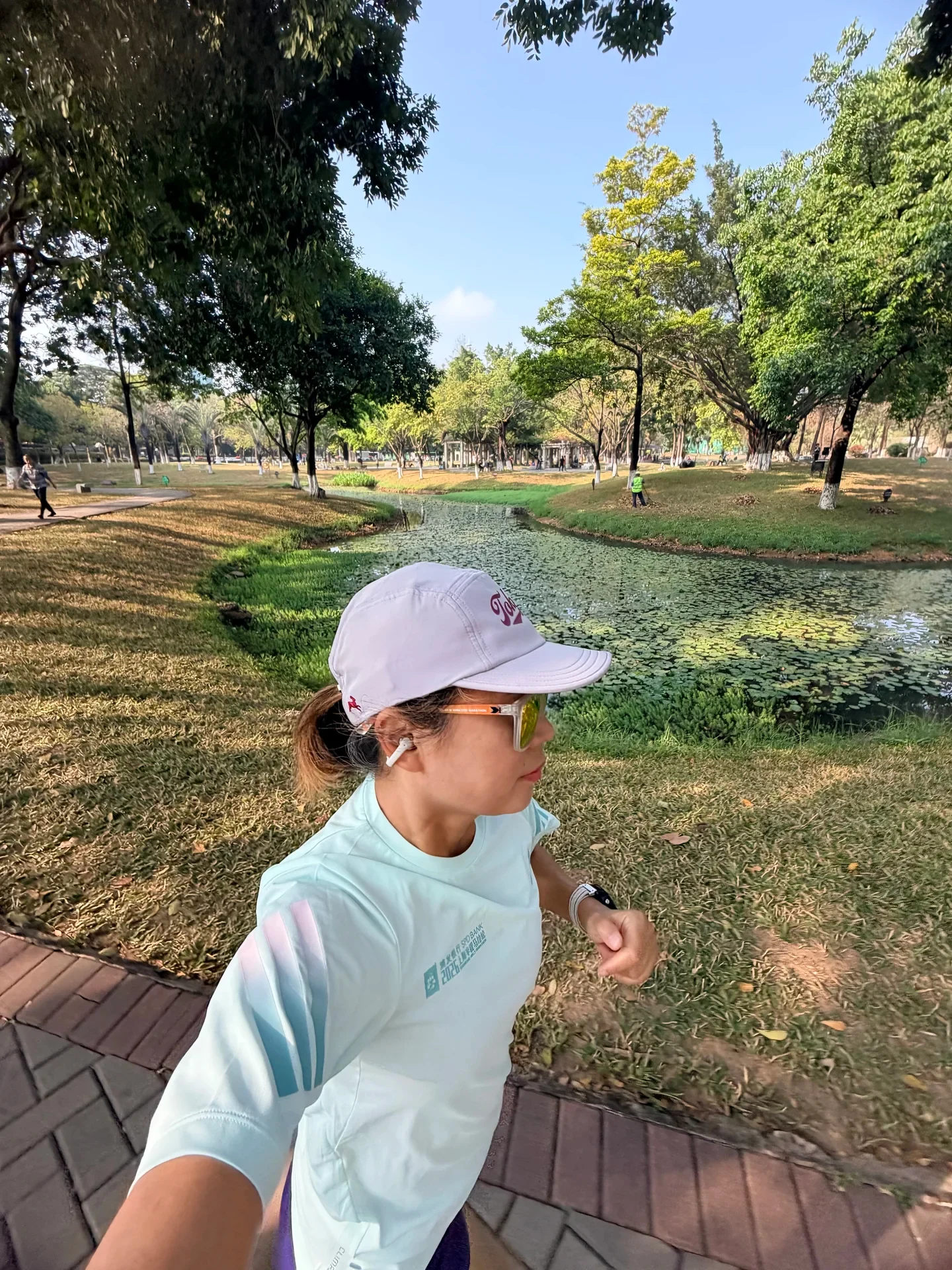 A woman runner in a white cap, sunglasses, and light blue t-shirt runs along a paved path in a sunlit park with a pond behind her, wearing a white clip-on earbud on her right ear.