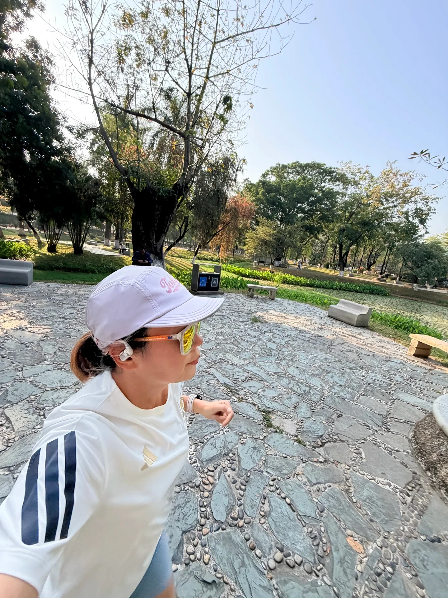 High-angle shot of a woman running along a stone-paved path in a park, wearing a white running cap, sunglasses, a white athletic shirt, and a white clip-on earbud on her right ear.