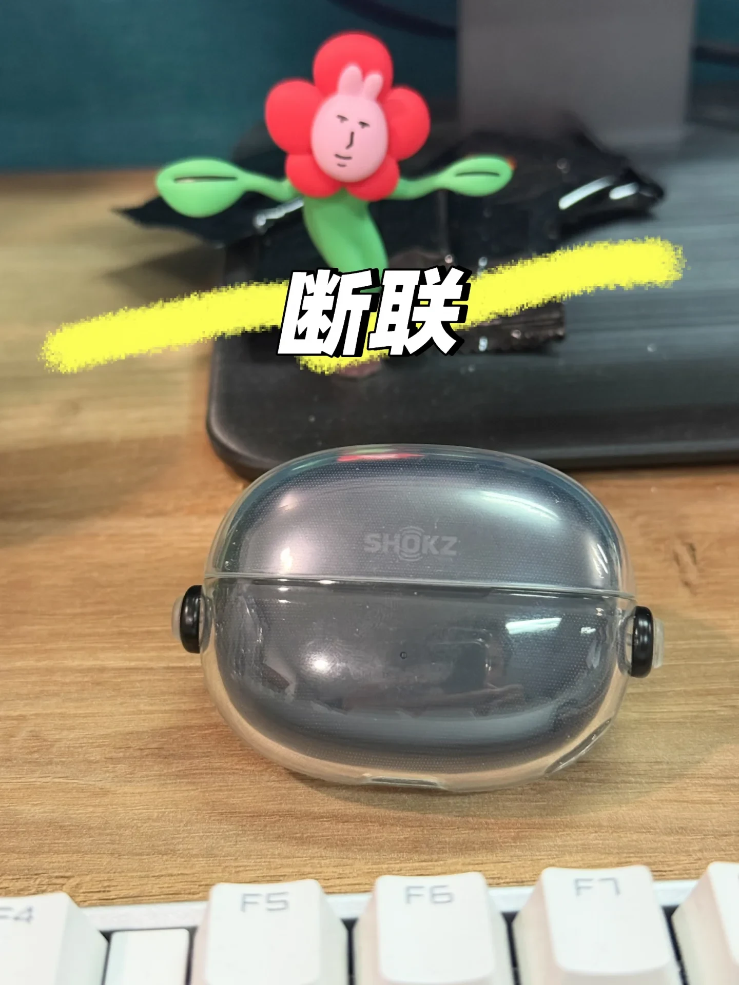 A dark gray Shokz earbud charging case inside a clear protective shell sits on a wooden desk in front of a keyboard, with a flower-shaped decoration and a black device behind it; a large Chinese text overlay reads “断联.”