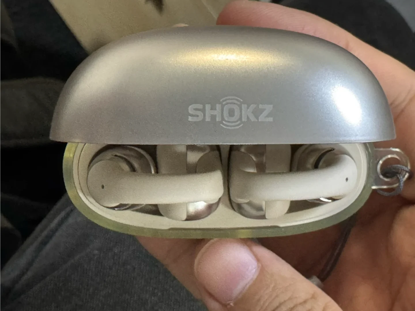 A hand holding the opened charging case of Shokz OpenDots earbuds in interstellar white, showing the two clip-on earphones nested inside and a clear protective case on the lower shell.
