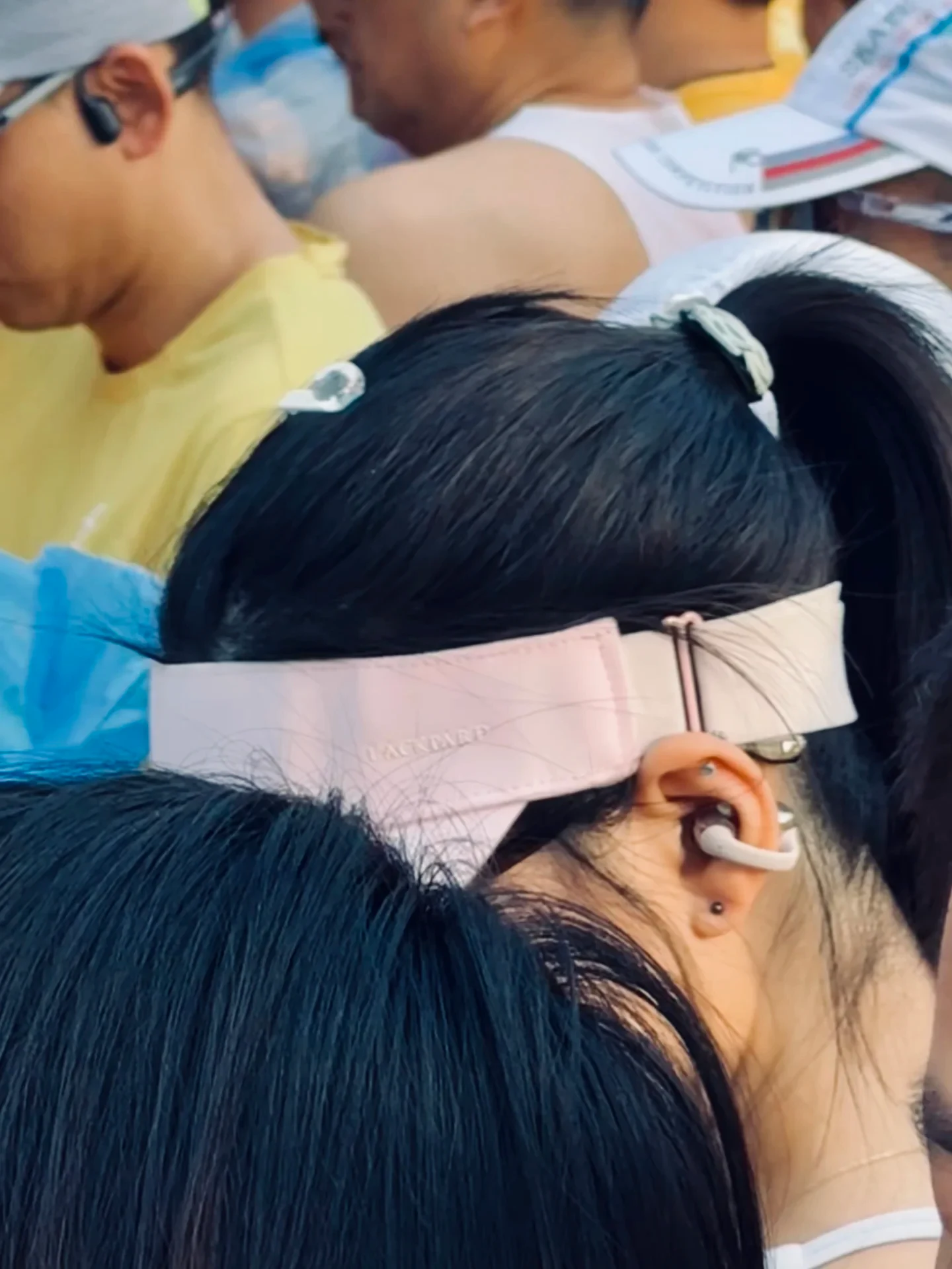 Rear side view of a person with dark hair tied back wearing a light pink running visor and a white clip-on earbud securely hooked onto the right ear amid an outdoor crowd.