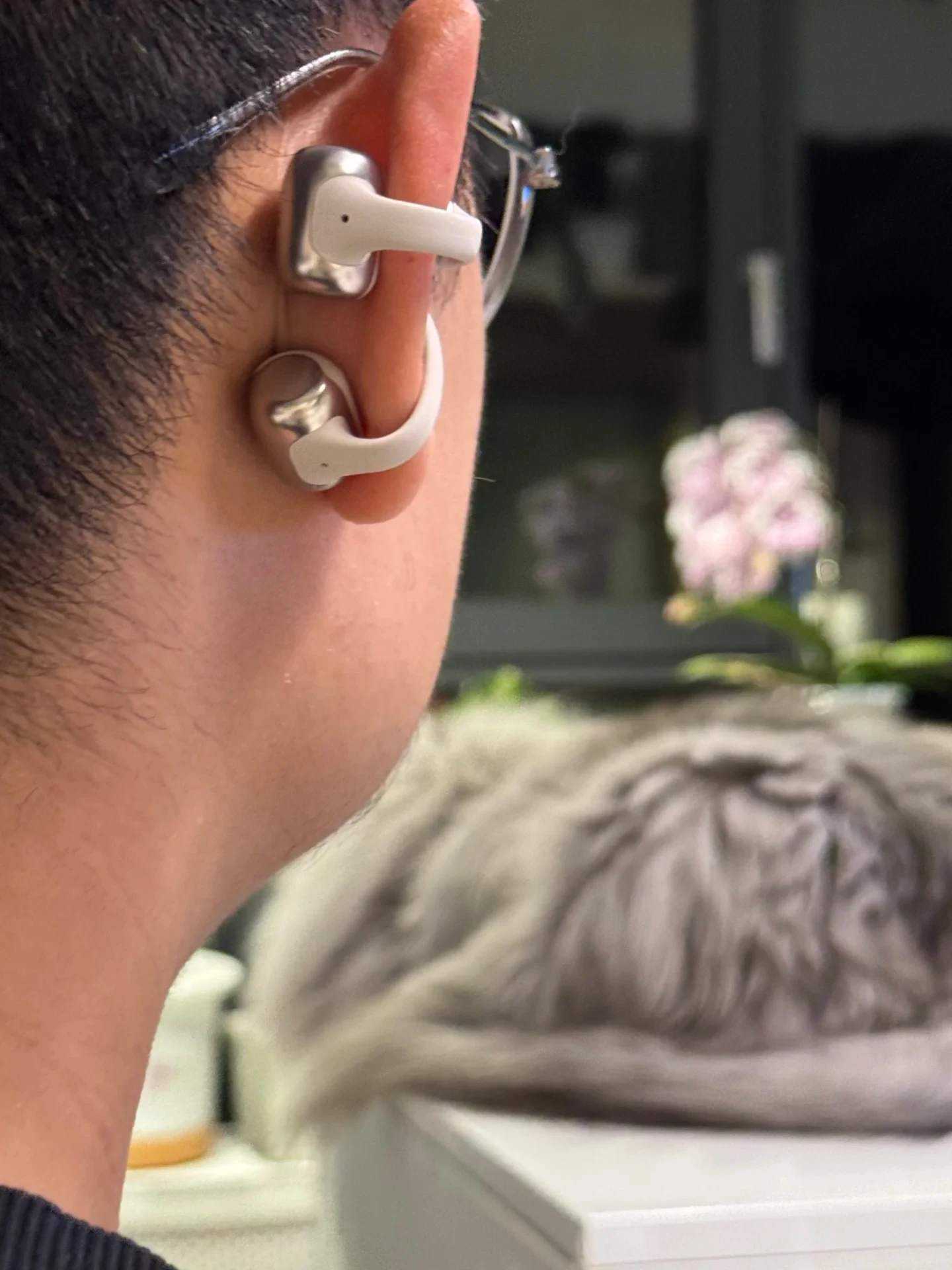 Close-up rear-side view of a person wearing glasses and a silver and white clip-on open-ear earbud on the outer ear.