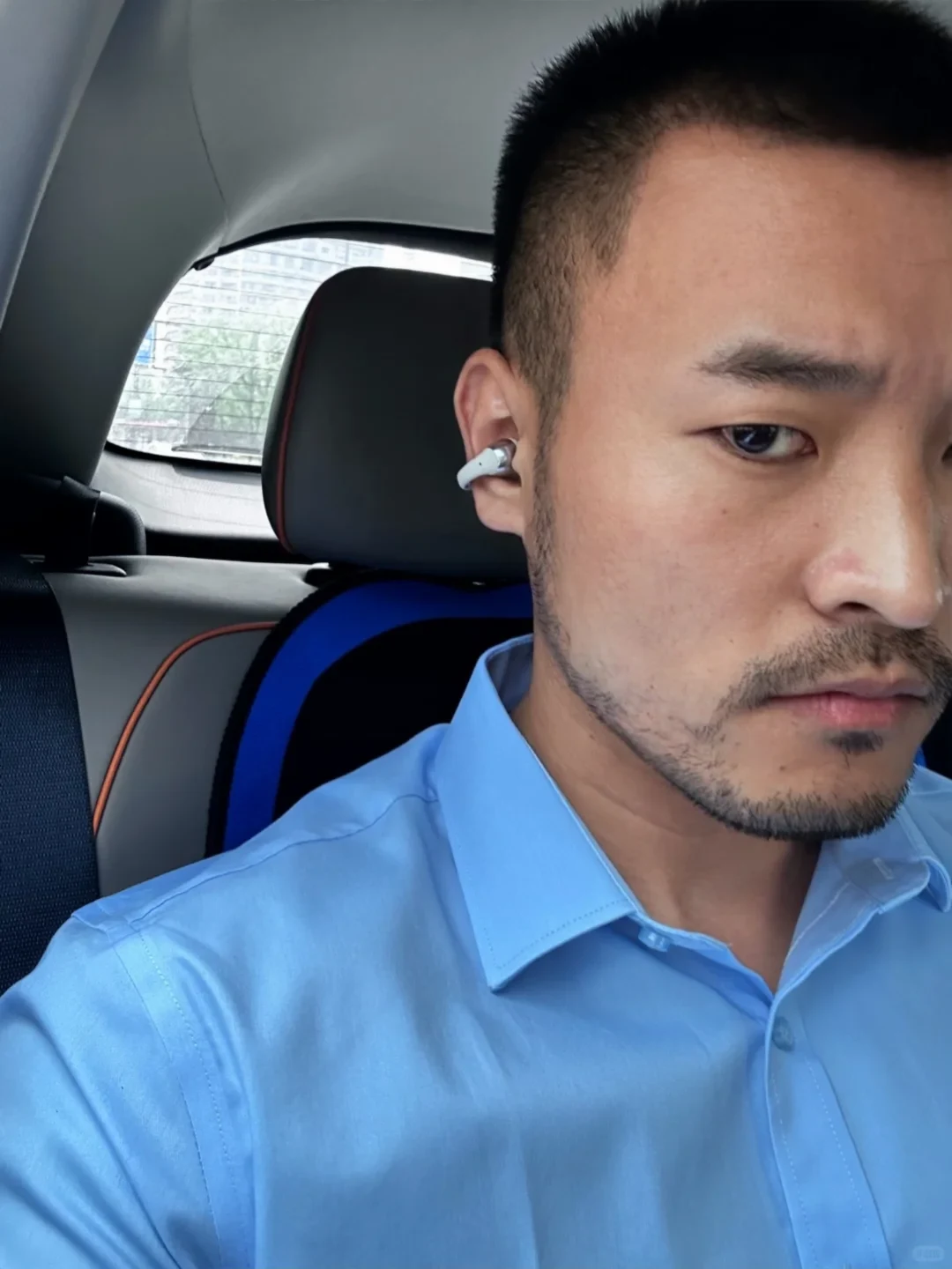 A close-up selfie of a man with short hair and light facial hair wearing a light blue dress shirt inside a vehicle. A small, clip-on style wireless earbud is visible in his right ear.