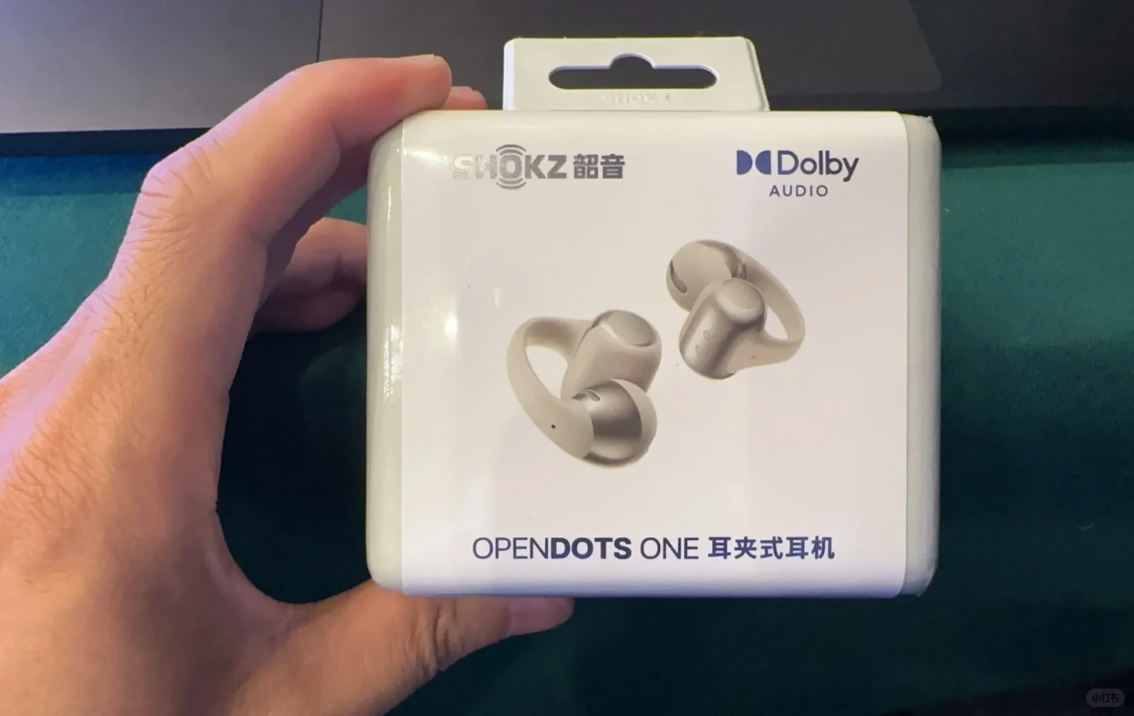 A hand holding the retail packaging of Shokz OpenDots One clip-on earphones, showing images of the beige open-ear earbuds and a Dolby Audio logo.