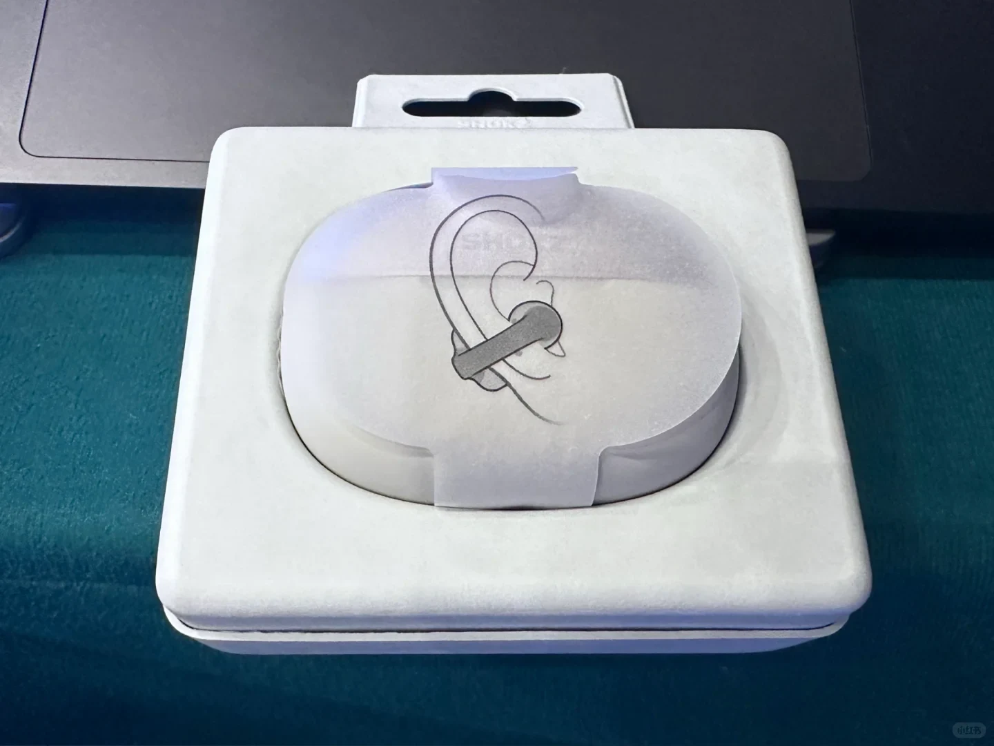 A white Shokz open-ear earphone charging case resting inside a molded white paper packaging tray. A translucent paper band wraps around the case, featuring an illustration showing how to wear the earbud hooked over the ear.