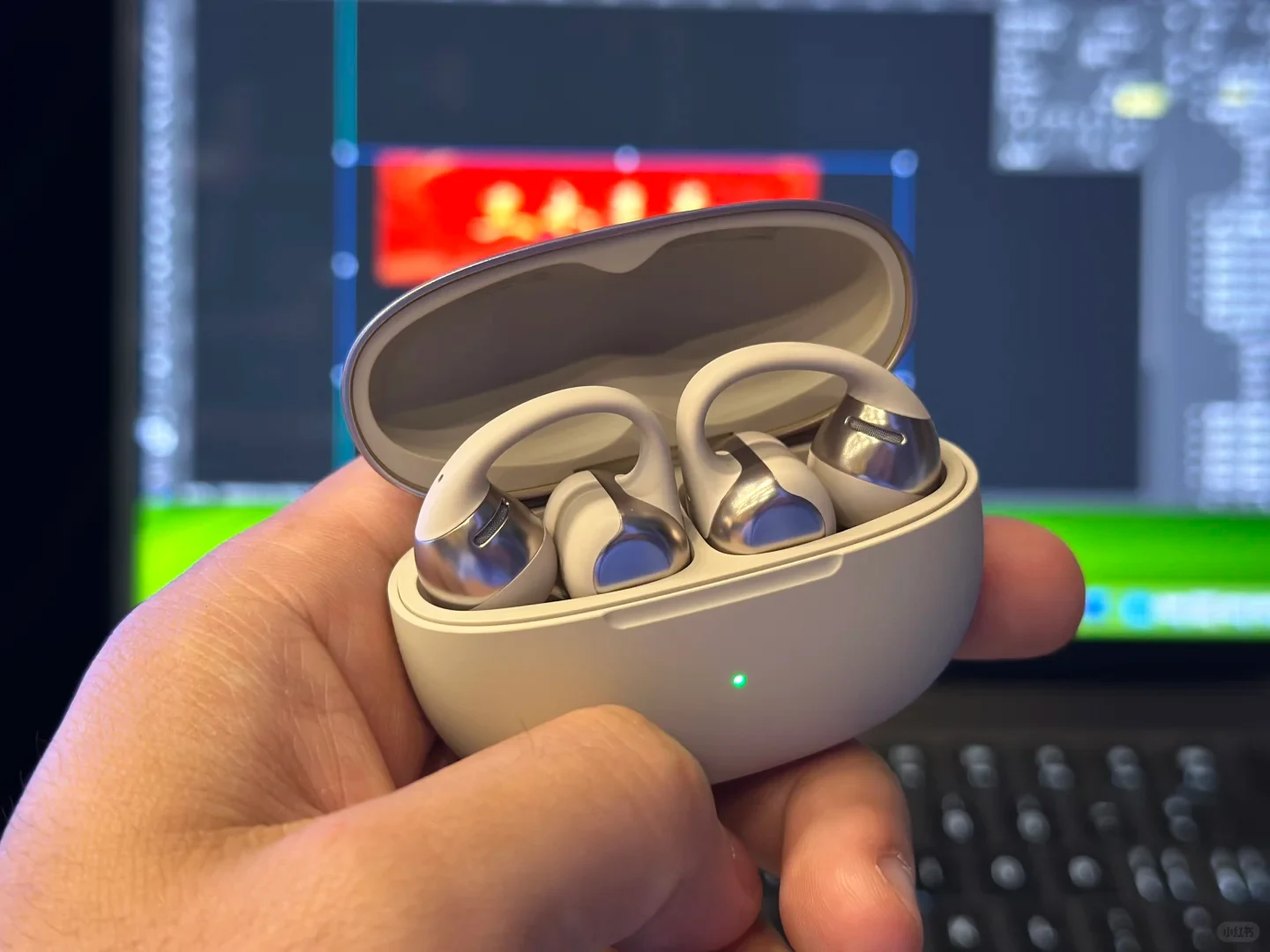 A close-up of a hand holding an open, off-white wireless earbud charging case. Inside, a pair of open-ear clip-style earbuds with metallic silver and matte beige finishes rest in their slots. A small green LED light illuminates on the front of the case, with a blurred computer monitor visible in the background.