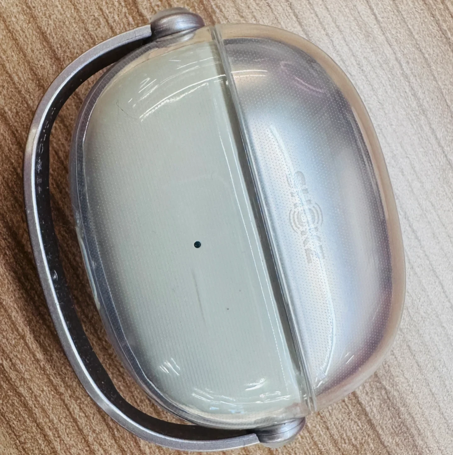 Top-down close-up of a Shokz OpenDots ONE charging case inside a clear protective cover with a black loop, resting on a wood-grain surface.