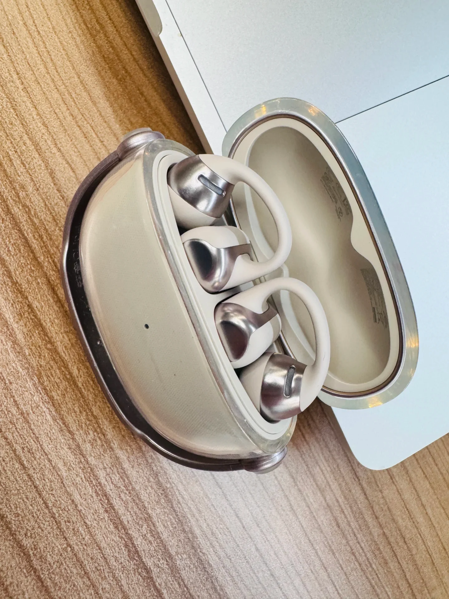 A pair of cream-colored and metallic Shokz OpenDots ONE clip-on earbuds sitting inside their opened charging case on a wooden tabletop next to a laptop.
