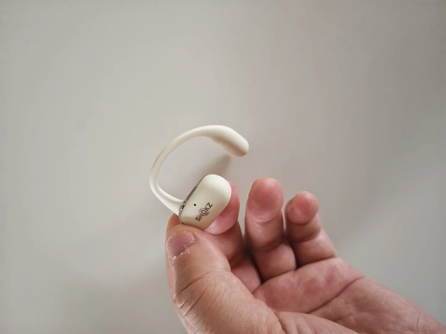 A hand holds an off-white Shokz open-ear earbud, showing its curved ear hook and speaker housing against a plain light background.
