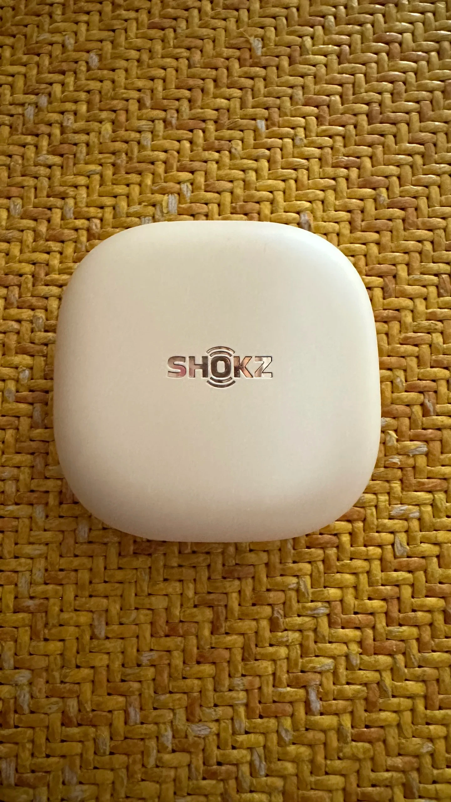 Top-down view of the rounded square Cloud White Shokz OpenFit 2+ charging case with a metallic Shokz logo centered on the lid, resting on a woven yellow textured surface.