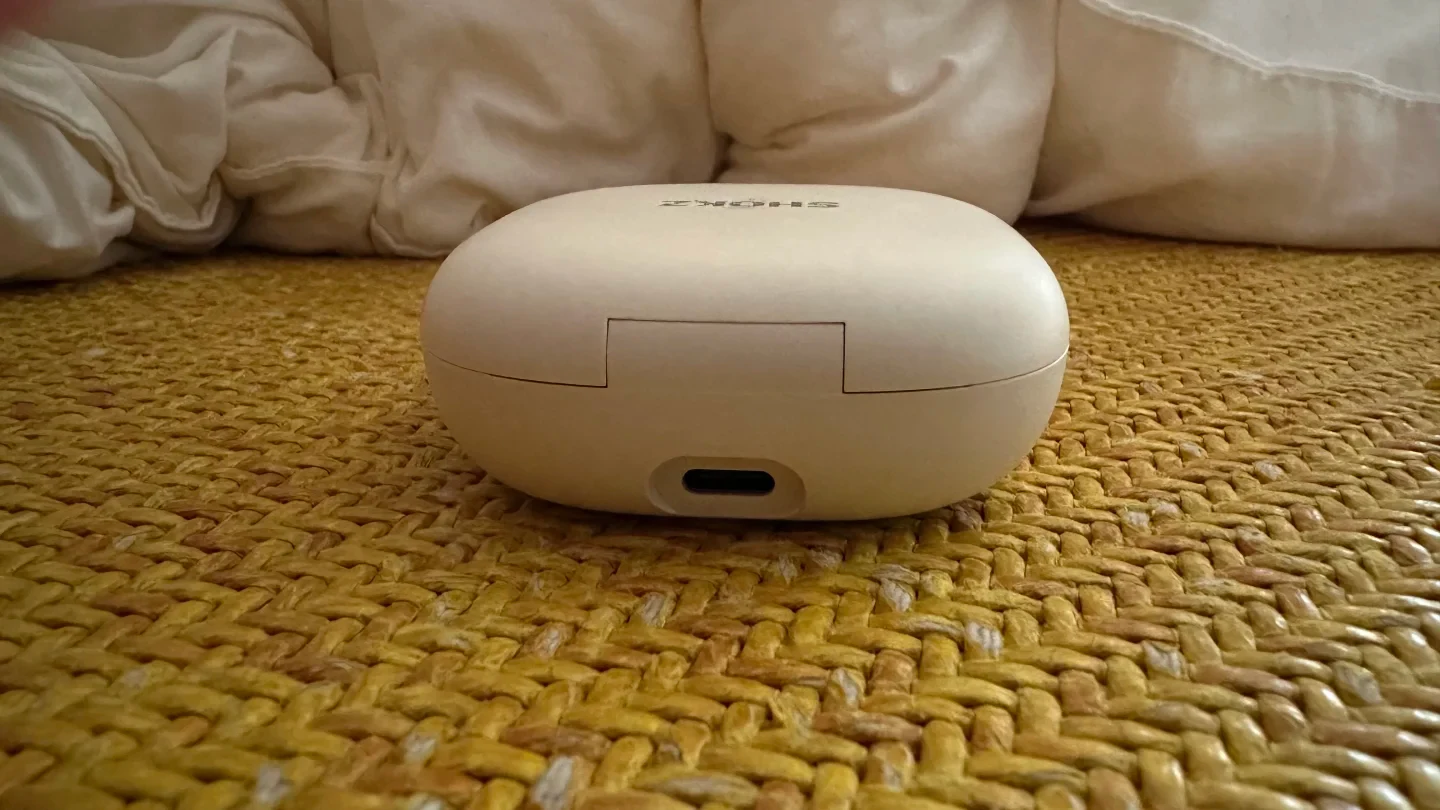 Rear view of the closed cloud white Shokz OpenFit 2+ earphone charging case showing its hinge and USB-C charging port on a woven yellow surface.