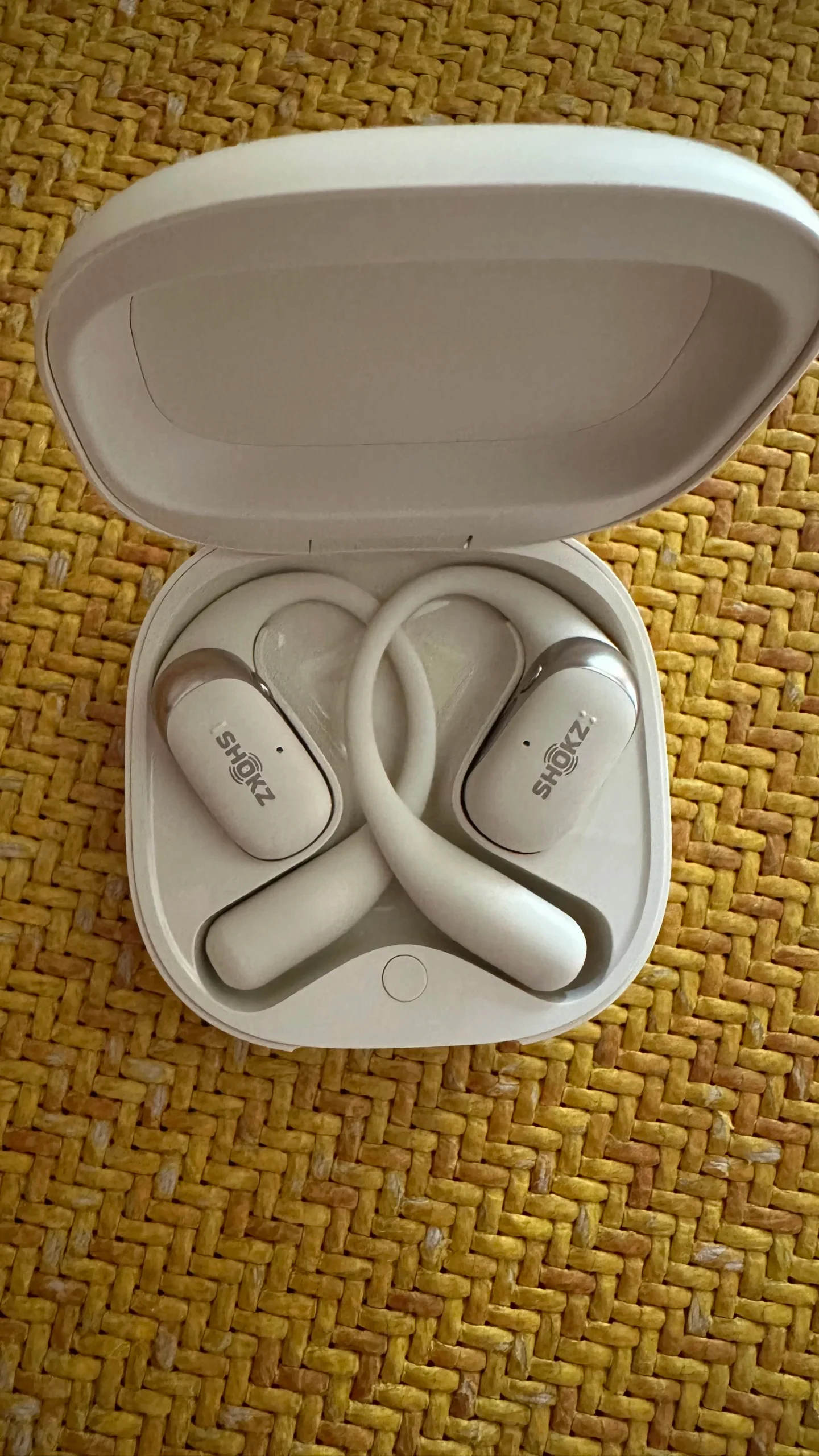 Top-down view of an open white charging case holding a pair of Shokz open-ear hook earbuds, placed on a woven yellow surface.