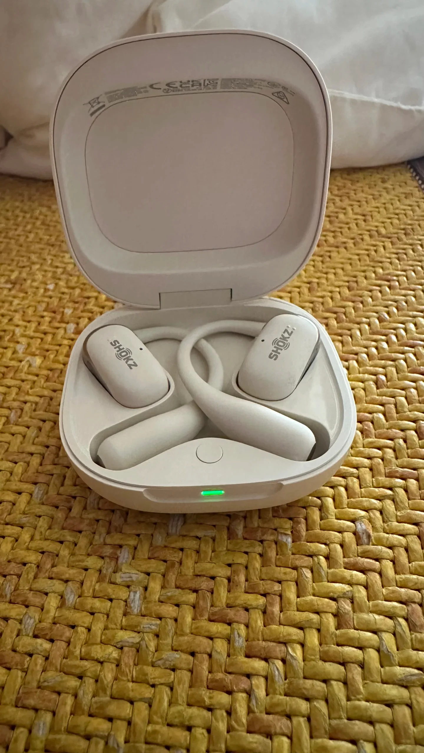 High-angle view of an open white charging case containing a pair of white Shokz OpenFit 2+ open-ear earbuds with a glowing green indicator light on a yellow woven surface.