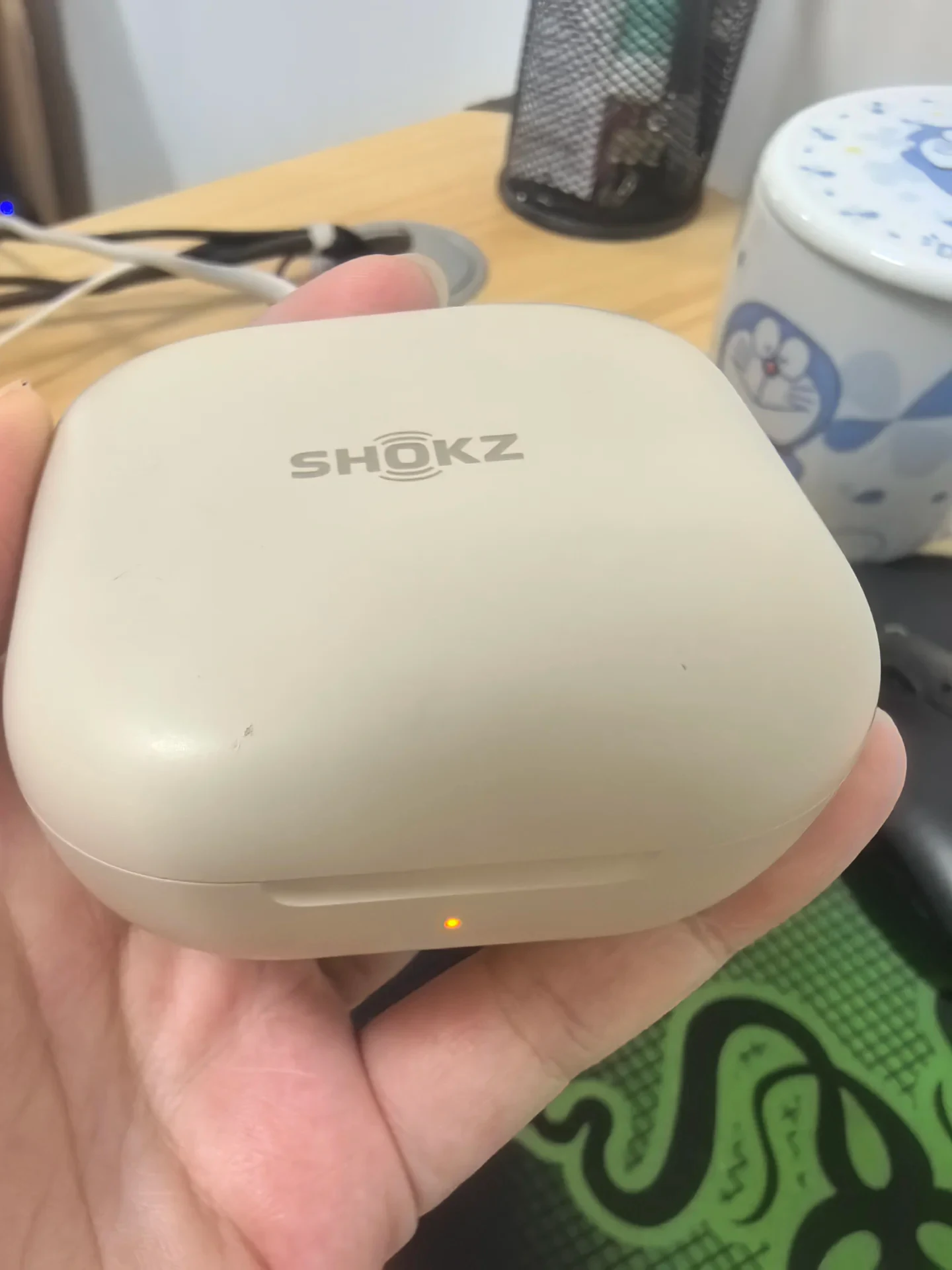 A hand holds a closed beige Shokz charging case with the SHOKZ logo visible and an amber indicator light illuminated on the front.