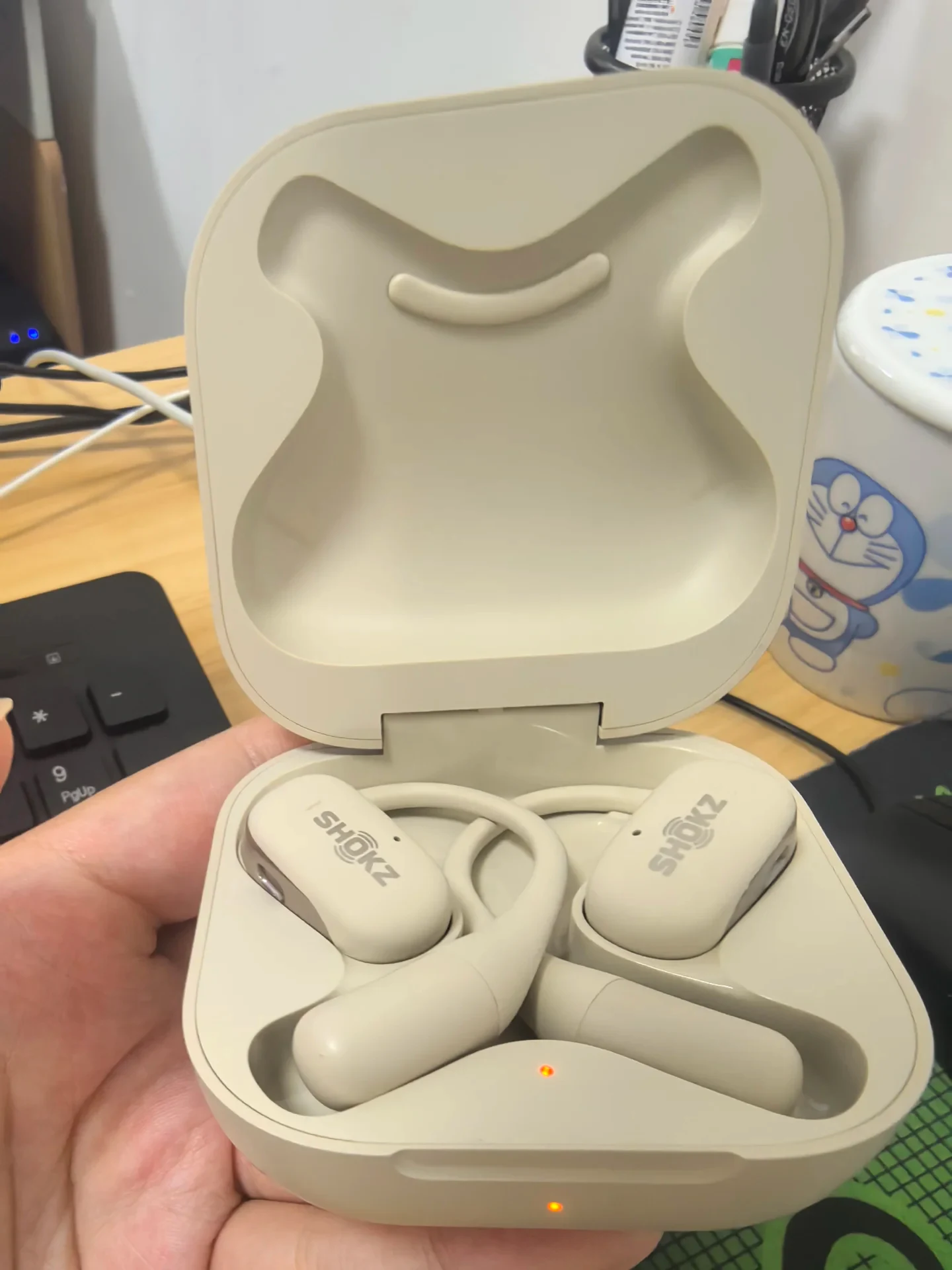 Open beige charging case held in a hand showing a pair of Shokz OpenFit open-ear earbuds resting inside with amber indicator LEDs illuminated.