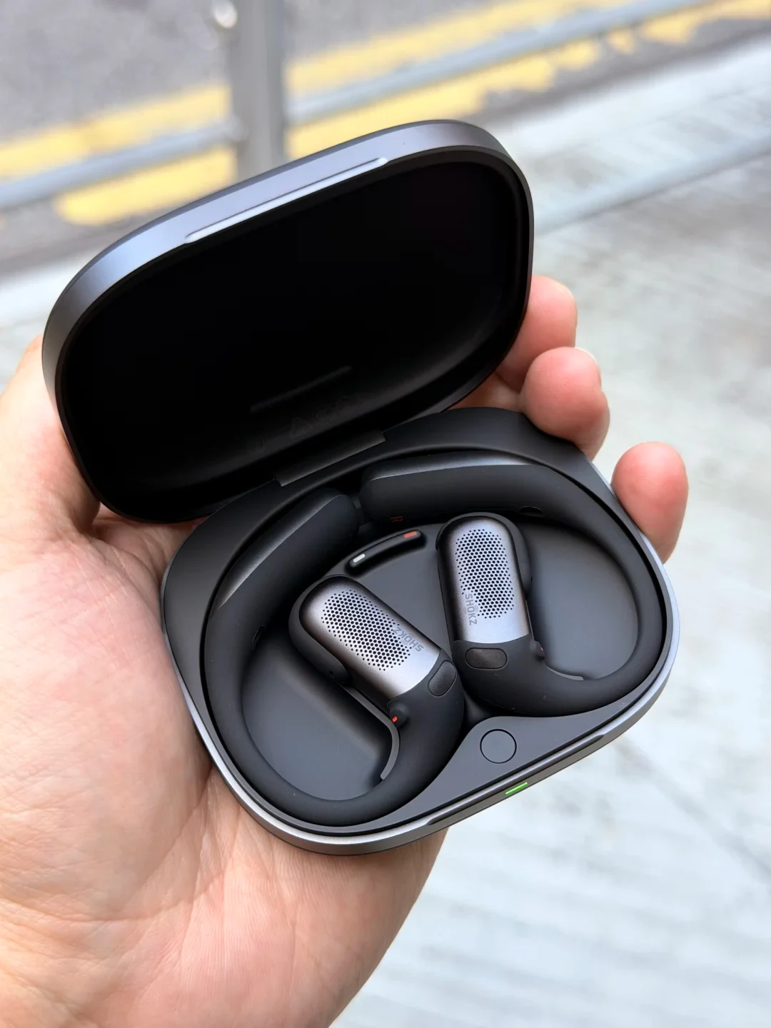 A hand holding an open dark gray charging case showing a pair of Shokz open-ear wireless earbuds nestled inside with curved black ear hooks and silver perforated speaker bodies.