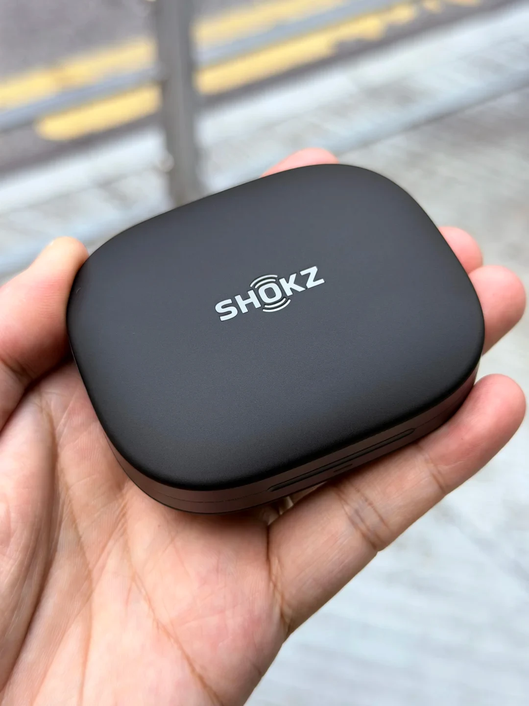 A close-up shot of a hand holding a matte black Shokz wireless earbud charging case outdoors.