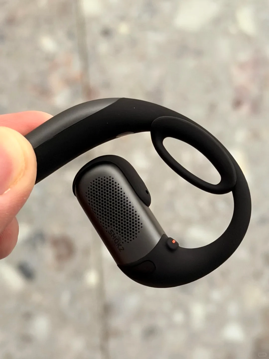 A close-up view of a black and dark metallic gray Shokz open-ear earbud held between fingers, featuring a curved earhook design, a perforated speaker grille with the "SHOKZ" logo, and a tiny orange indicator light.