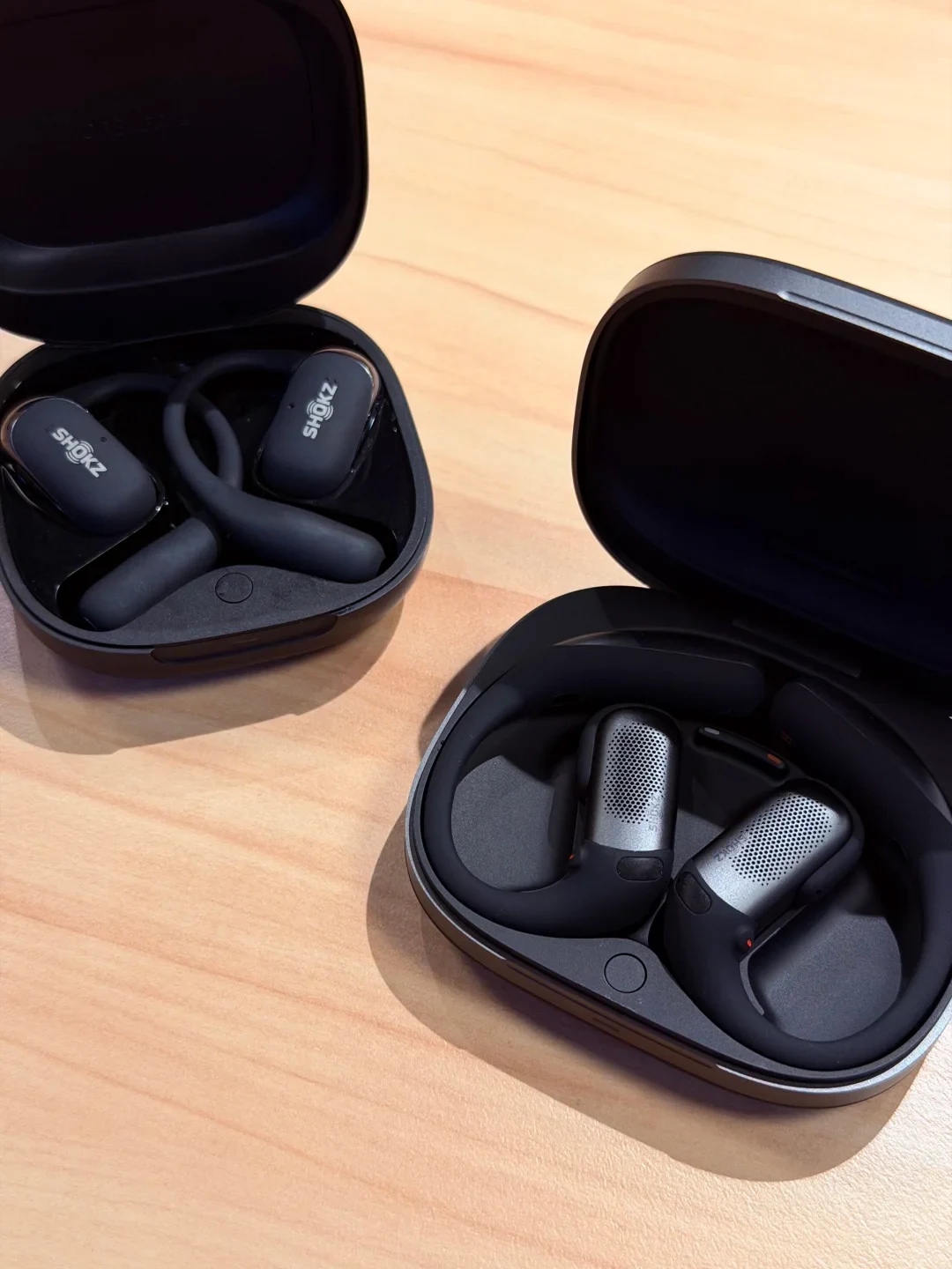 Two open charging cases displaying Shokz open-ear wireless earbuds side by side on a light wooden surface. The case on the upper left contains solid black earbuds, while the case on the lower right contains earbuds with textured silver speaker grilles.