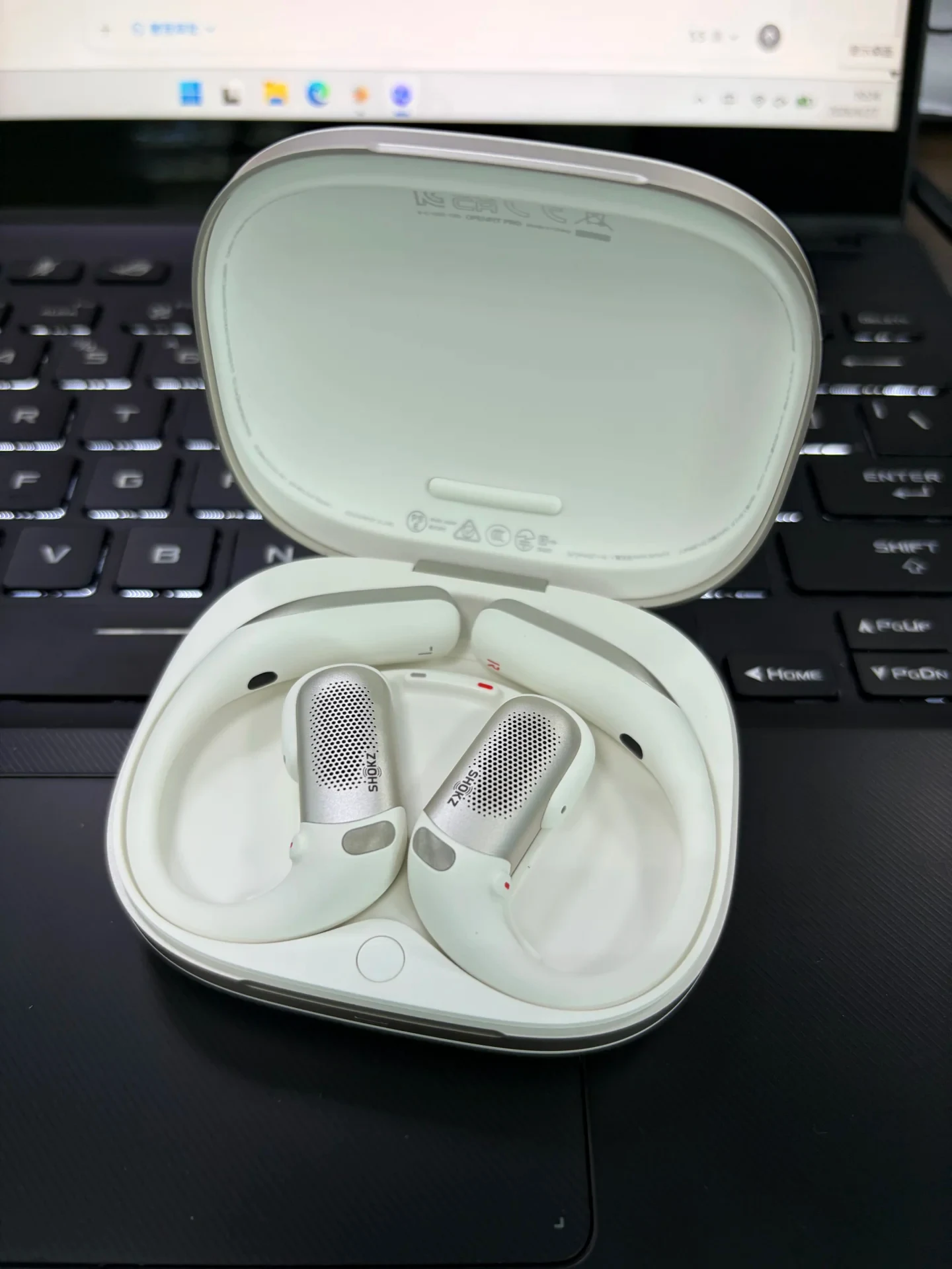High-angle close-up of a pair of beige Shokz OpenFit Pro open-ear earbuds seated inside their open charging case on a laptop keyboard.