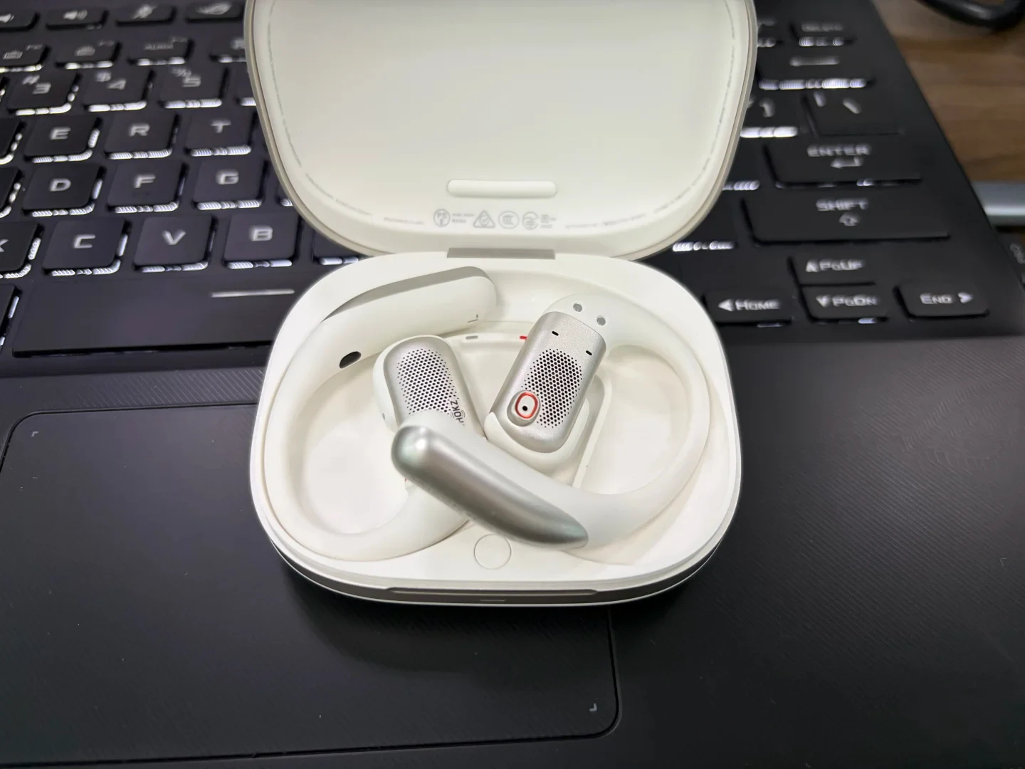 An open white charging case holding a pair of white and silver open-ear hook earbuds, resting on a black laptop keyboard and touchpad.