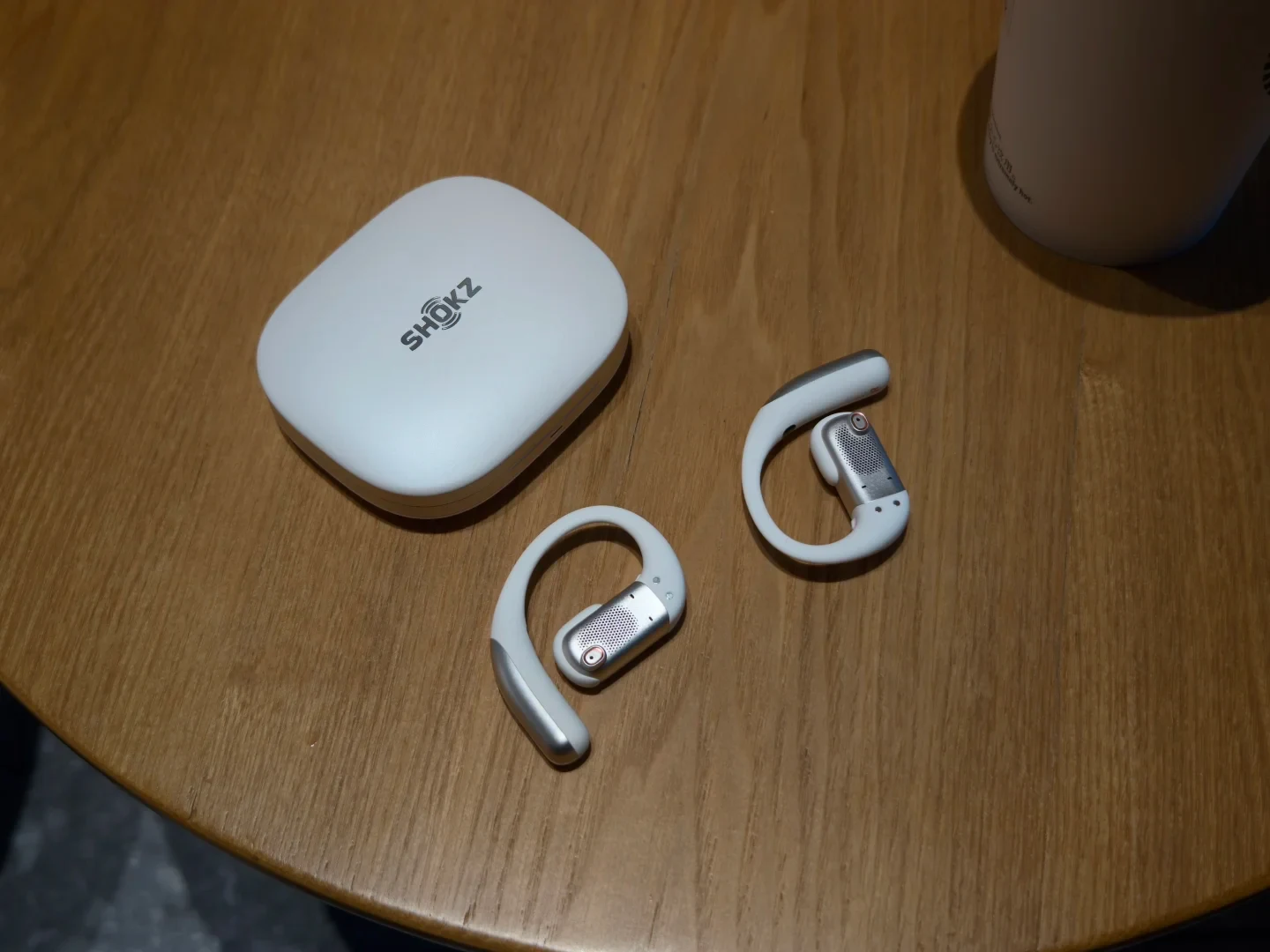 A pair of white and silver open-ear earbuds lying on a wooden table next to their rounded white charging case bearing the Shokz logo.
