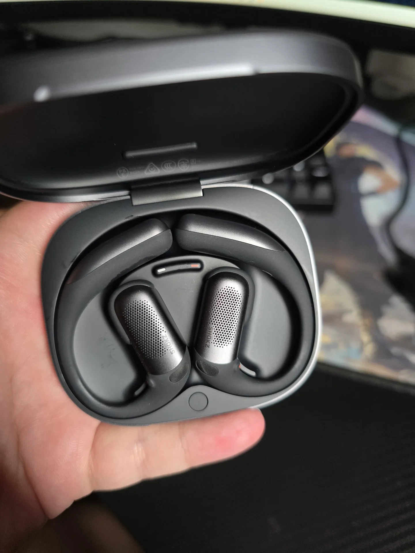 Open charging case held in a hand, displaying a pair of dark gray Shokz OpenFit Pro open-ear hook earbuds seated inside.