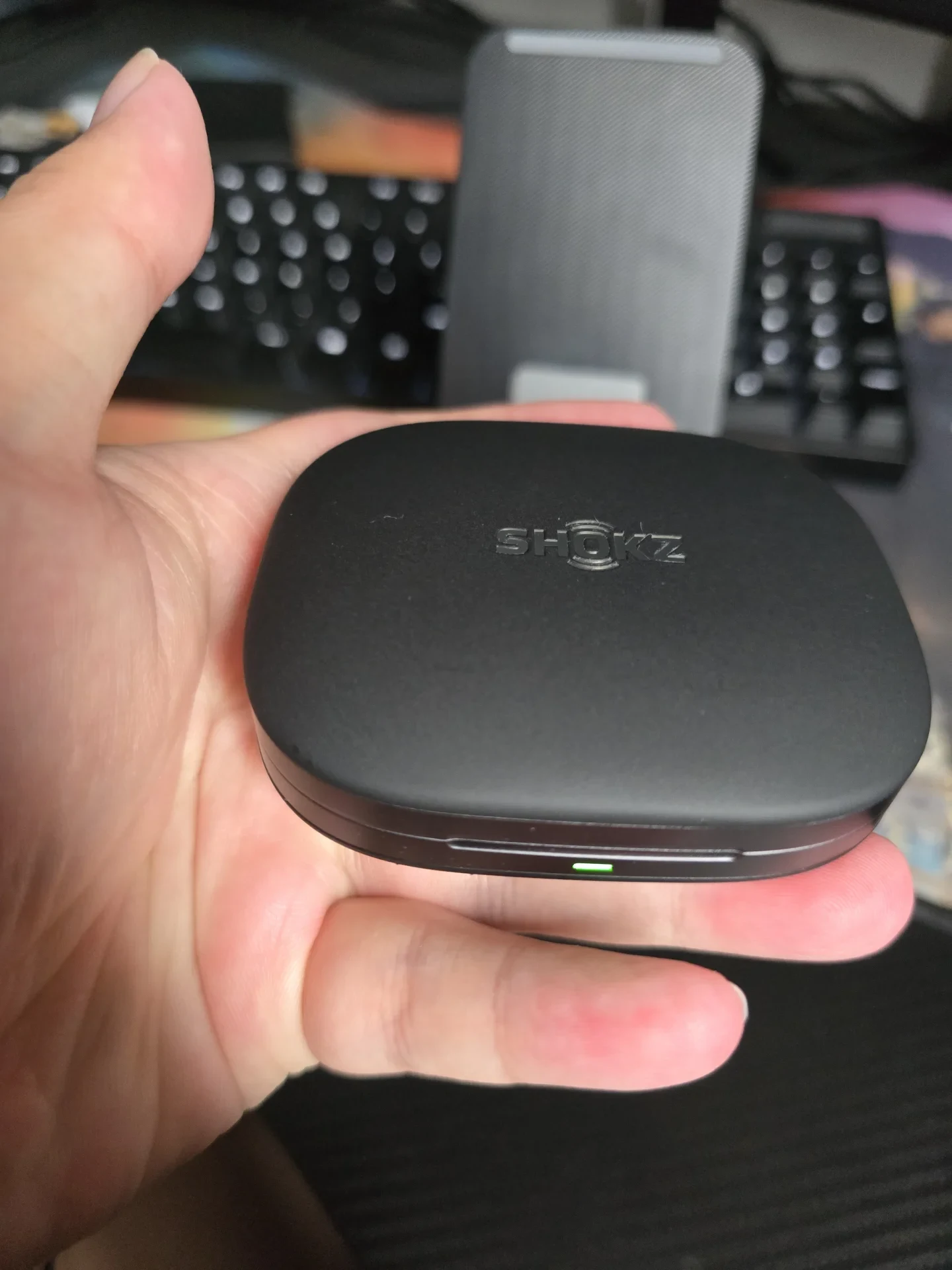 A hand holding the closed matte black charging case of Shokz open-ear headphones, displaying the debossed SHOKZ logo on the top lid and an illuminated green indicator light on the front rim.