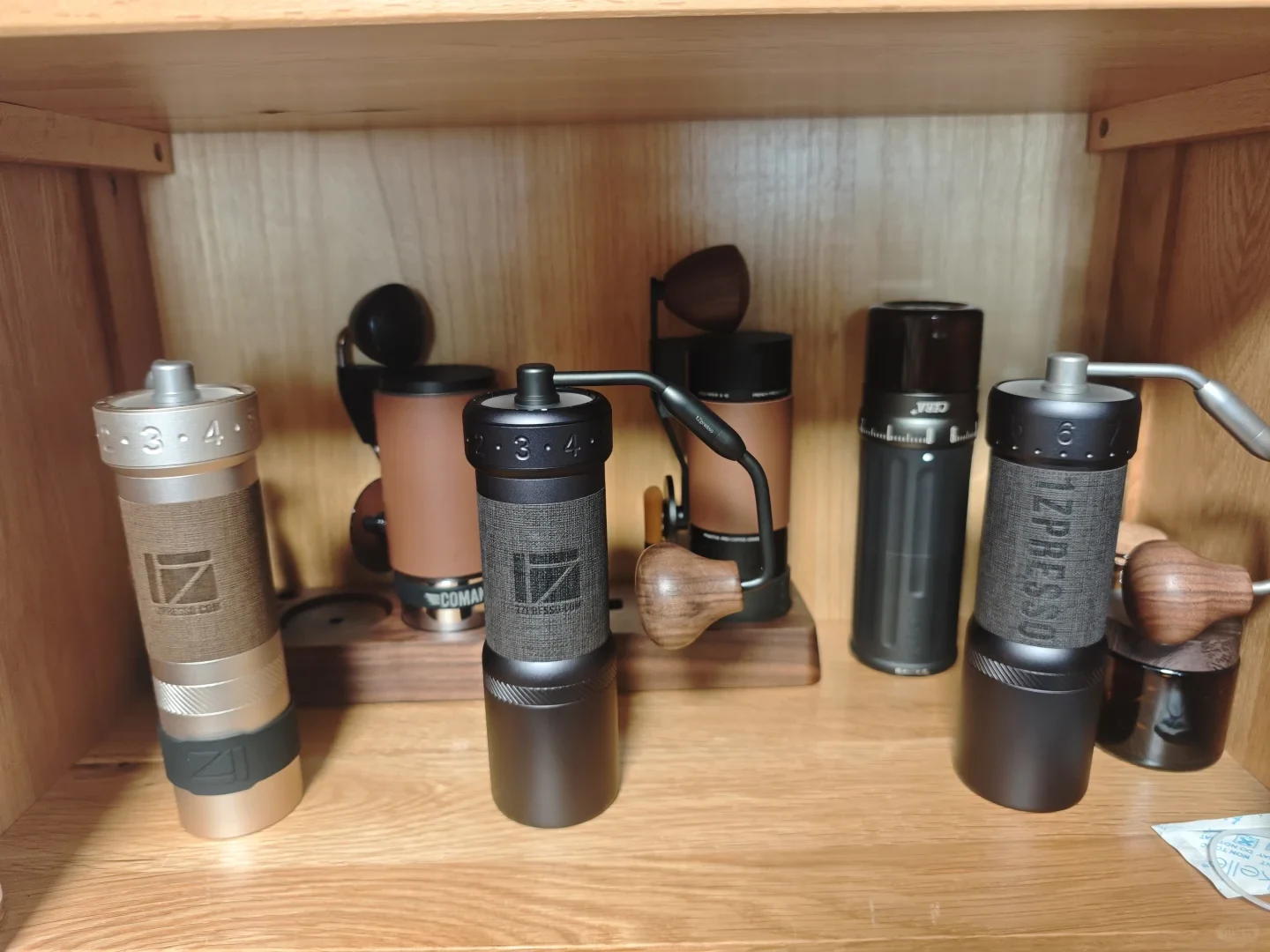 Six coffee grinders stand on a wooden shelf: three 1Zpresso hand grinders in front, with Comandante, MAVO and CERA grinders behind them.