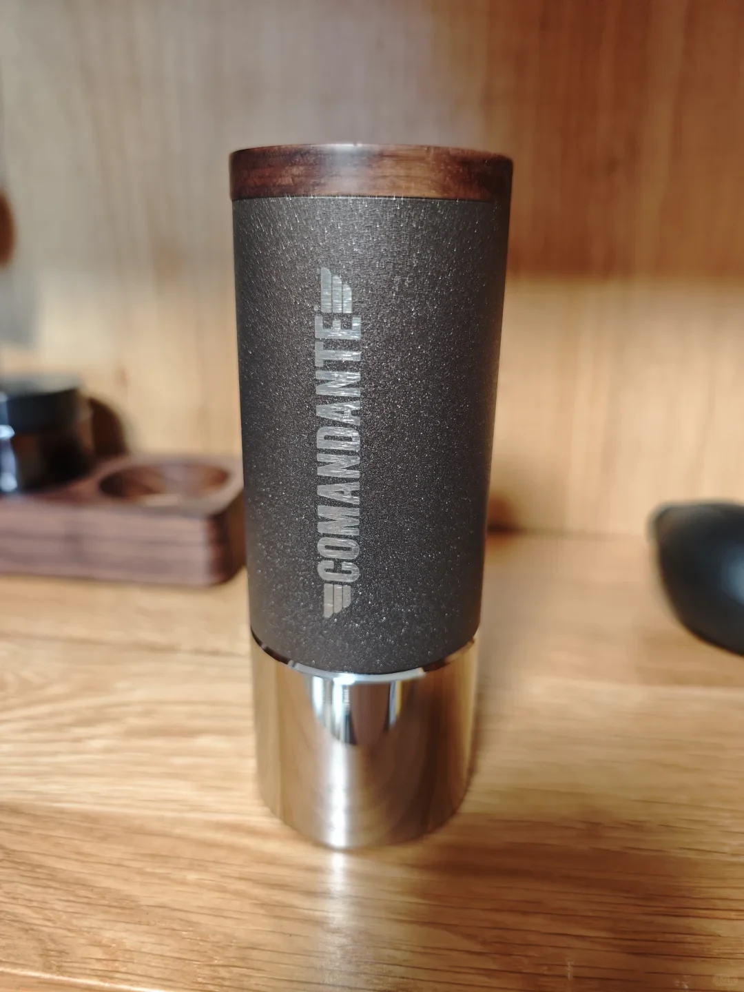 A Comandante grinder body with a dark textured sleeve, wooden lid and metal grounds container stands without its crank on a wooden table.