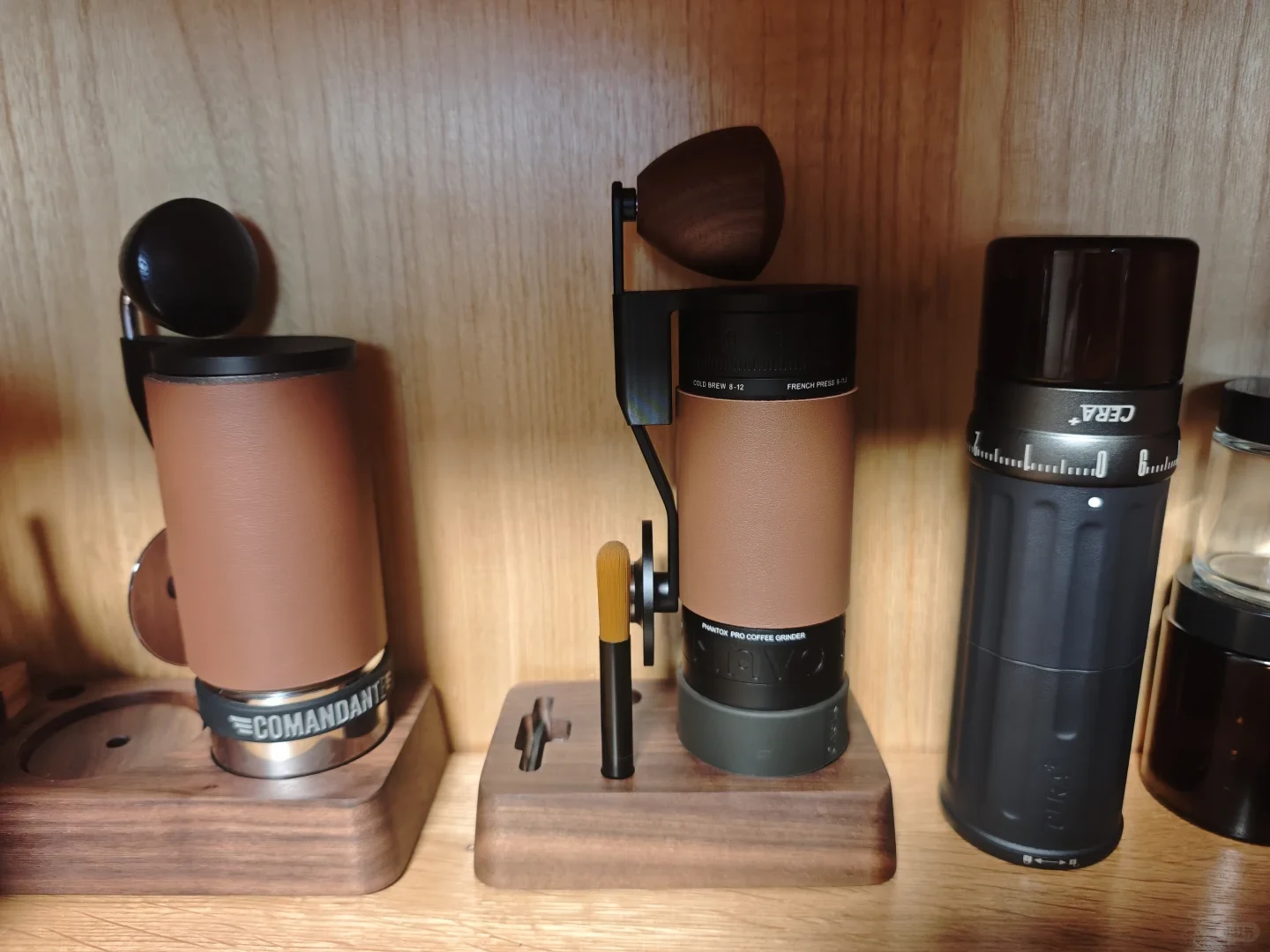 Coffee grinders and brewing equipment displayed on wooden shelves, including two brown-and-black hand grinders, a black grinder with a numbered adjustment ring, and partially visible glass coffee equipment at the right.