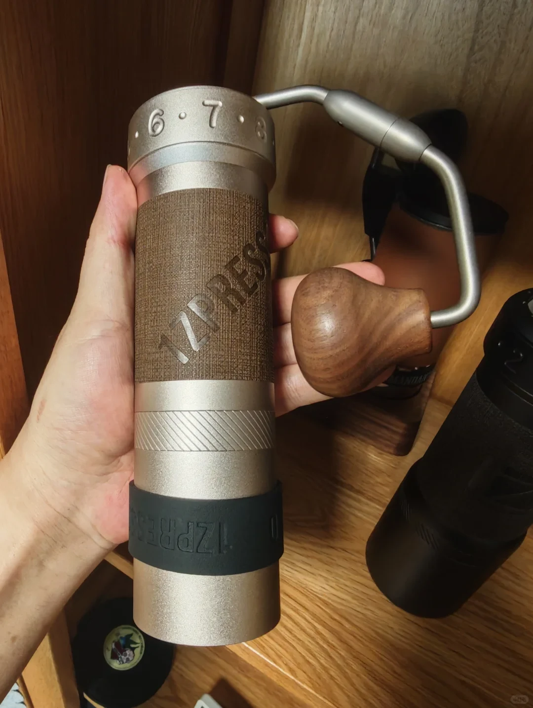A hand holds a metal manual coffee grinder with a wooden crank handle; other dark-colored grinders are visible on the wooden shelf behind it.
