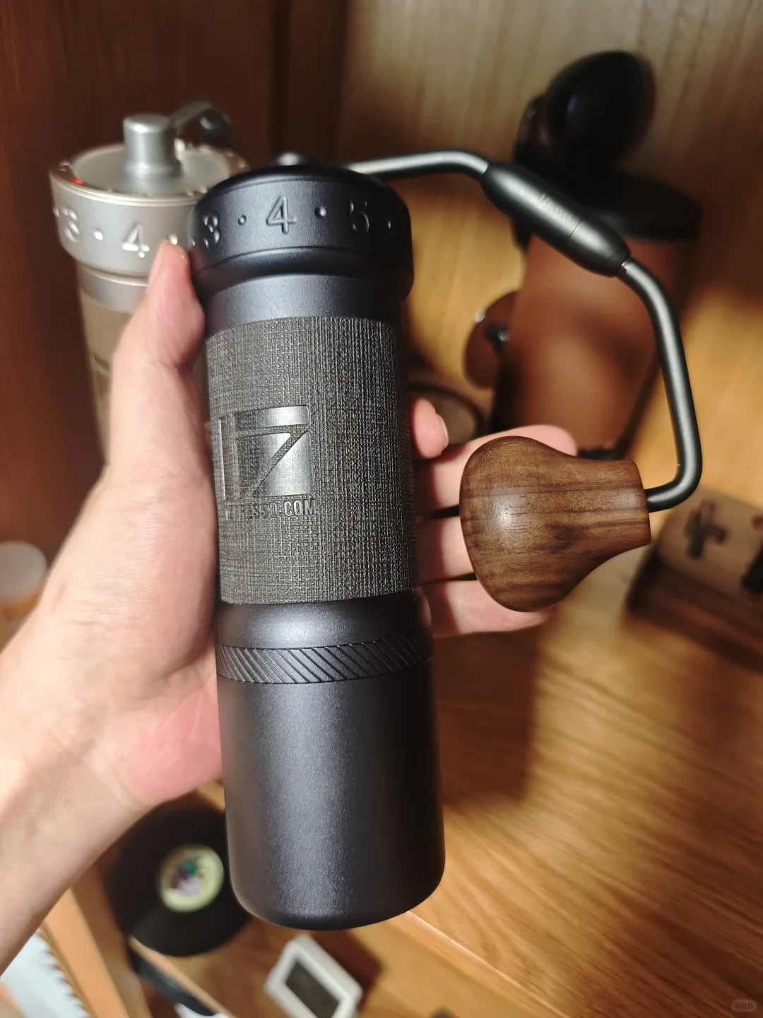 A hand holds a black manual coffee grinder with a wooden crank knob; another metal grinder and coffee equipment sit on a wooden surface behind it.