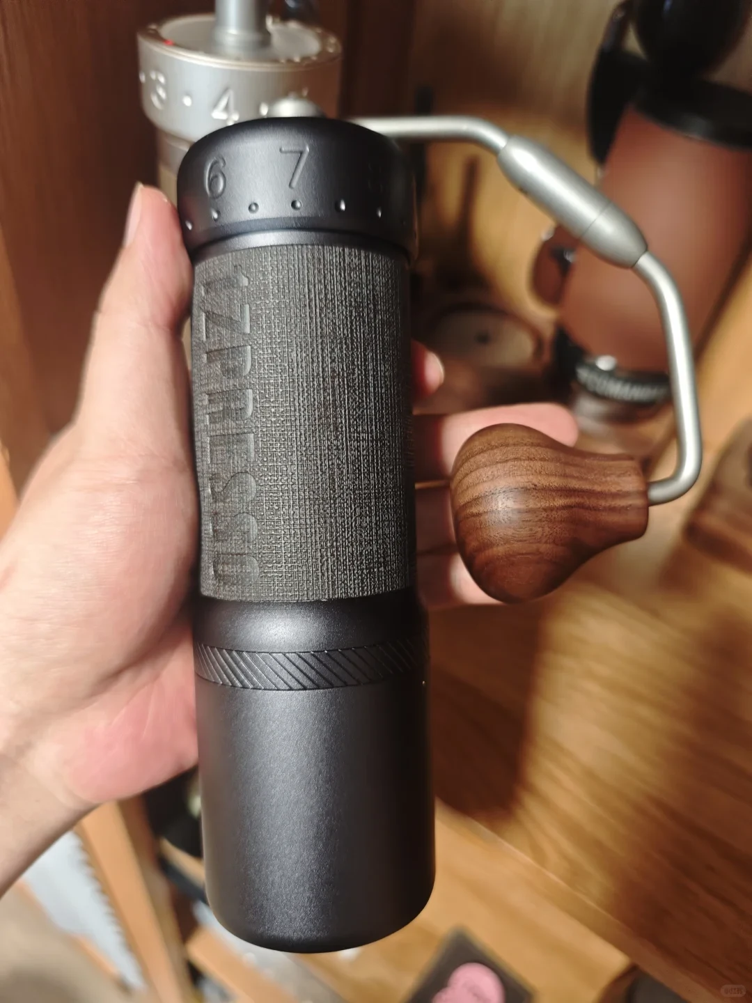 A hand holds a black manual coffee grinder with a textured grip, numbered adjustment dial, and metal crank ending in a wooden knob; coffee equipment sits on a wooden counter behind it.