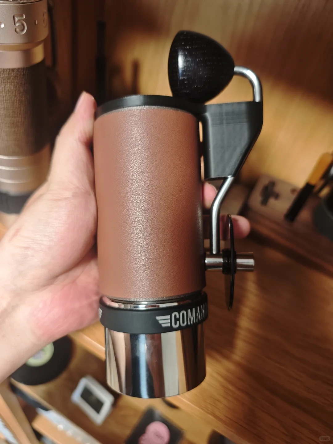 A hand holds a brown-and-black manual coffee grinder with a side crank and a metal grounds container, above a wooden counter.