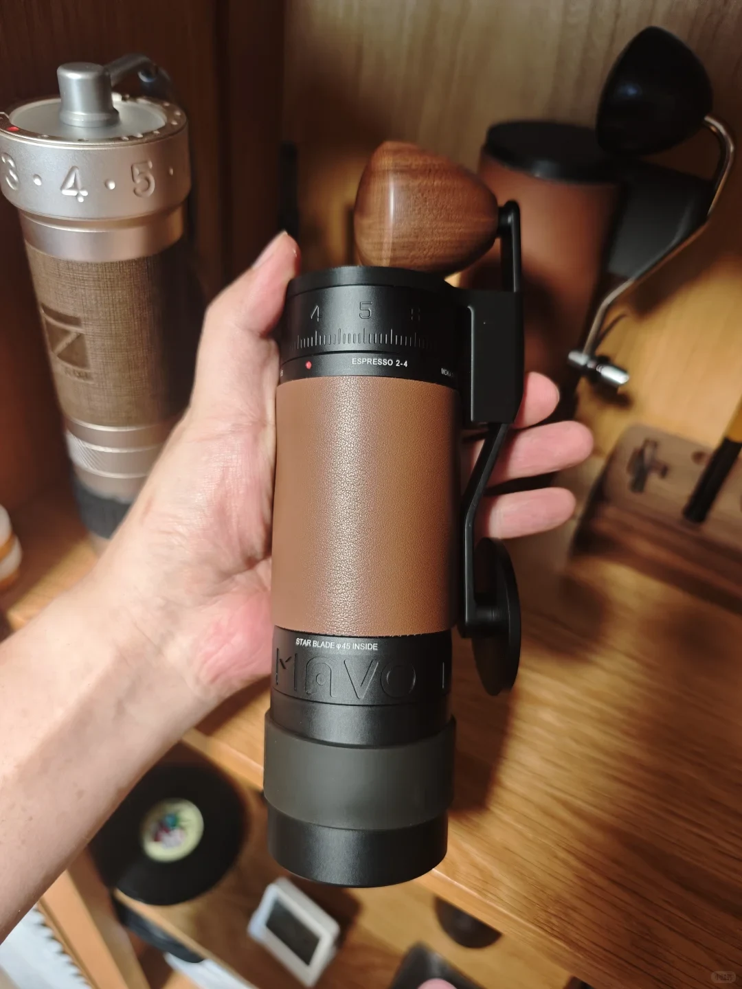 A hand holds a brown-and-black manual coffee grinder with a wooden crank handle and numbered grind-setting dial, in front of other coffee equipment on a wooden counter.