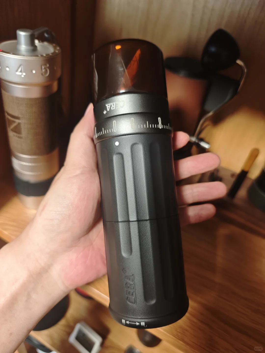 A hand holds an inverted black CERA+ portable electric coffee grinder with a translucent grounds jar and a numbered adjustment ring.