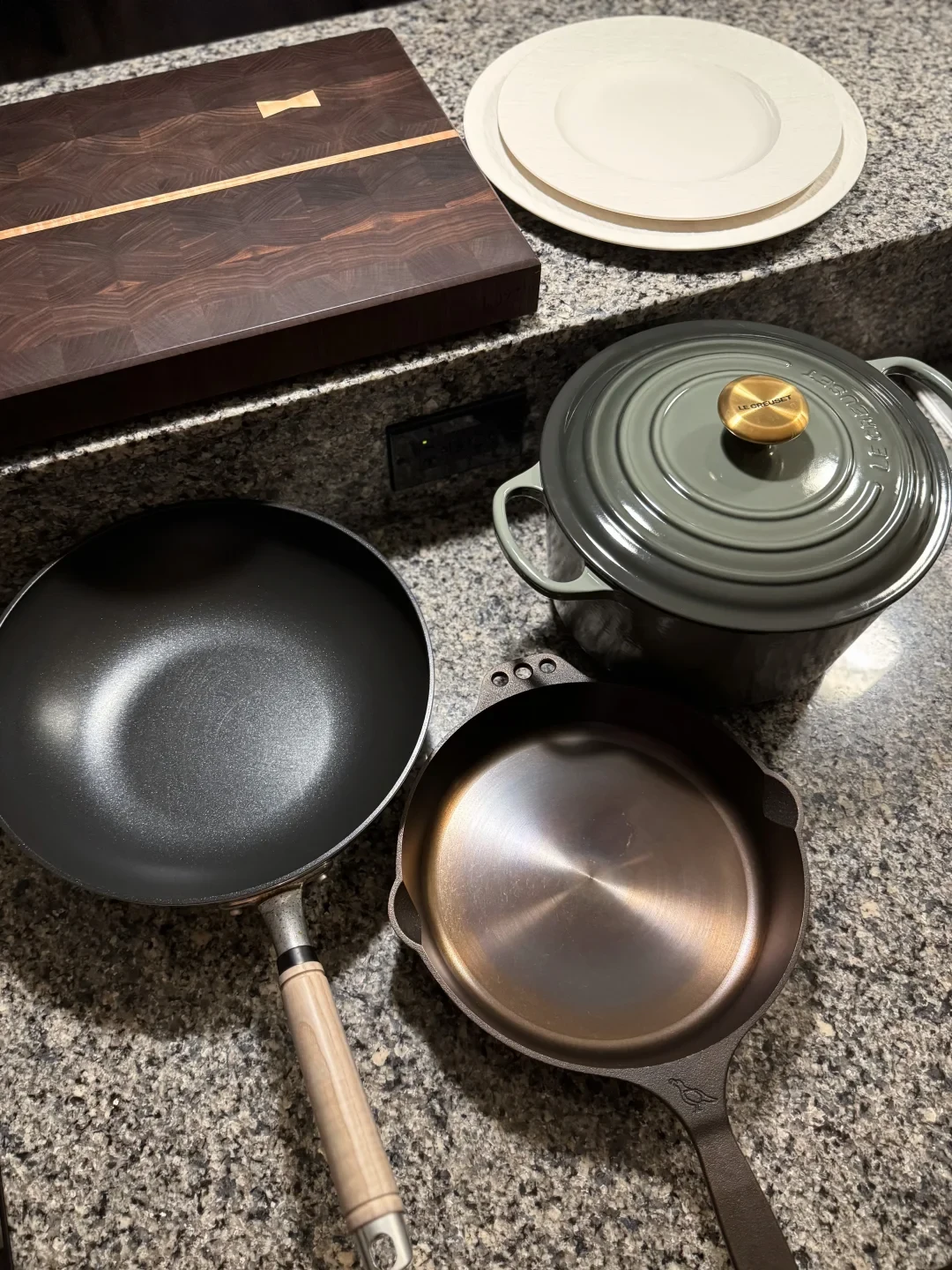 A kitchen countertop holds a dark wooden cutting board, stacked white plates, a lidded gray pot, a black wok with a wooden handle, and a bronze-colored skillet.