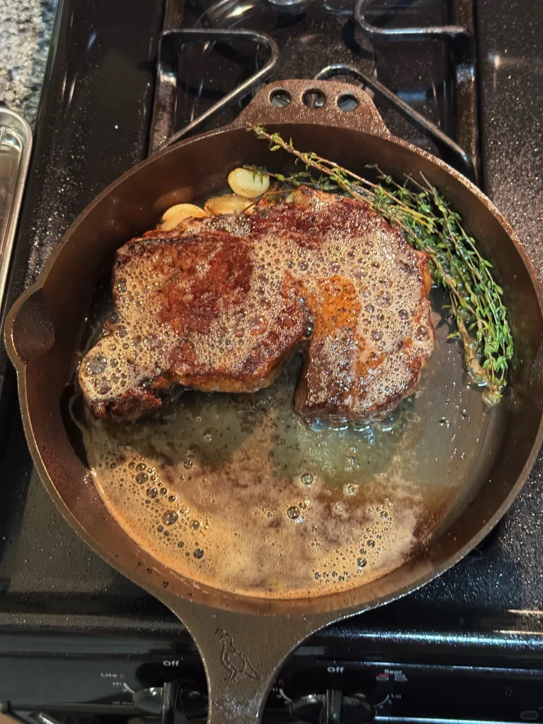 Two browned steaks sear in foaming butter with garlic and herbs in a frying pan on a stovetop.