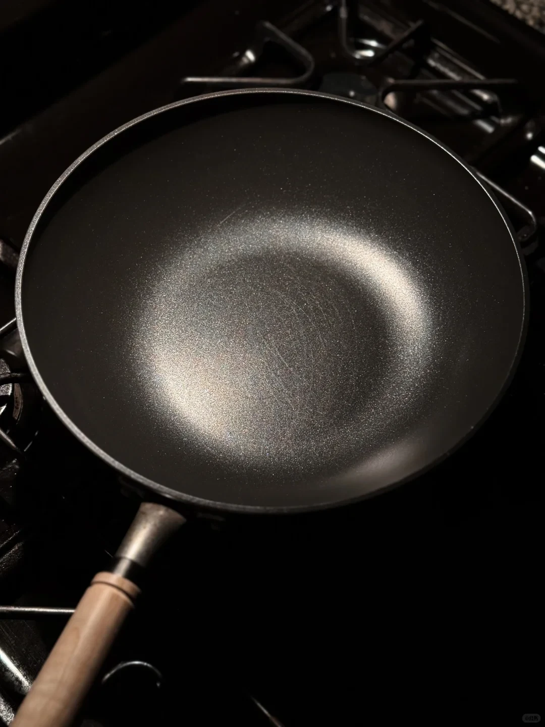 A dark, empty wok with a speckled interior and light wooden handle rests on a black gas stovetop.
