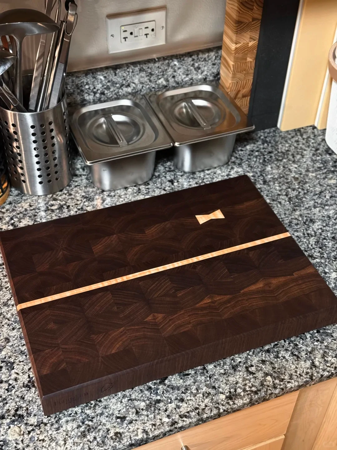 A thick, dark end-grain wooden cutting board with a narrow light-colored inlay across it sits on a granite kitchen counter. Two lidded stainless-steel containers and a utensil holder are behind it.