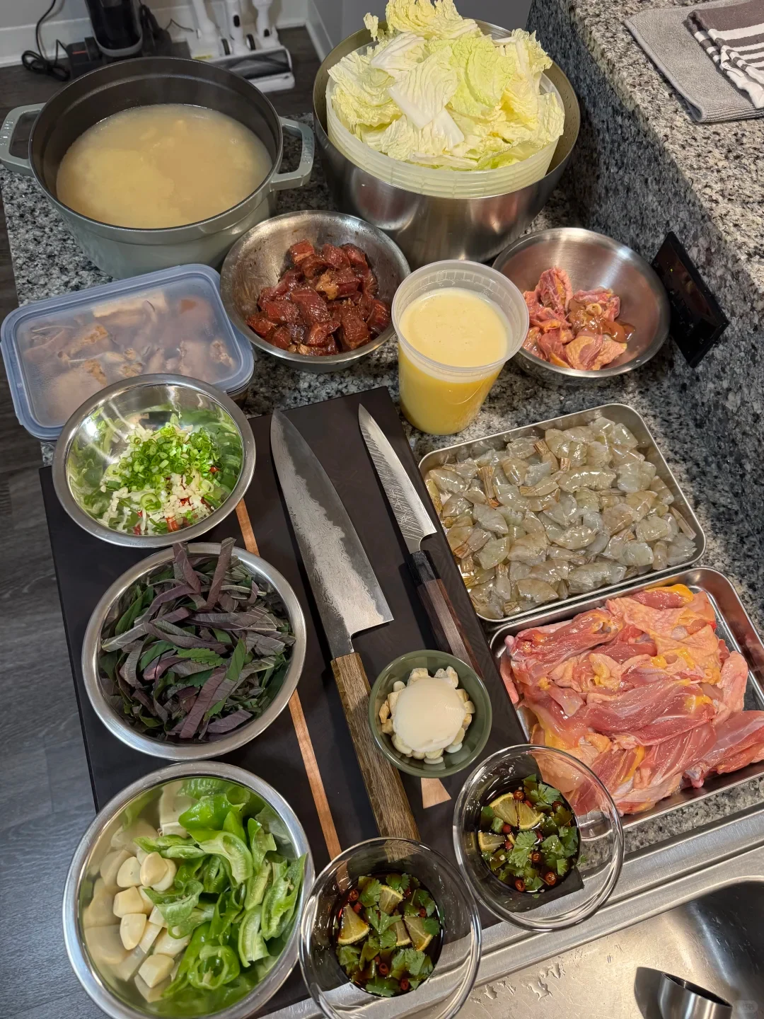 An overhead kitchen-prep photo shows bowls and trays of chopped vegetables, leafy greens, shrimp, raw chicken, and other ingredients arranged around two knives on a cutting board, with a pot of broth and a large bowl of cabbage at the back.