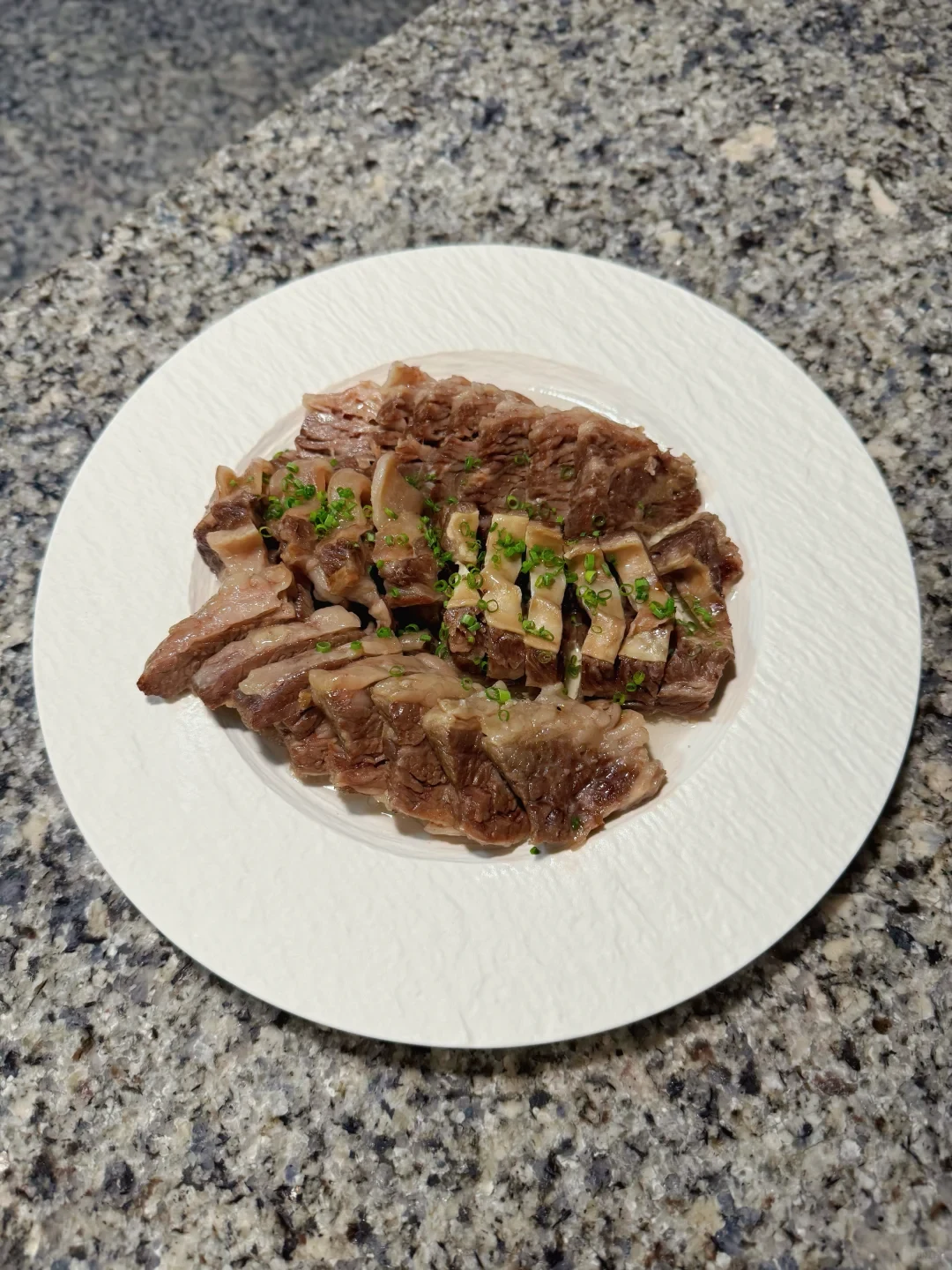 A white plate of sliced cooked meat topped with chopped green herbs sits on a speckled stone countertop.