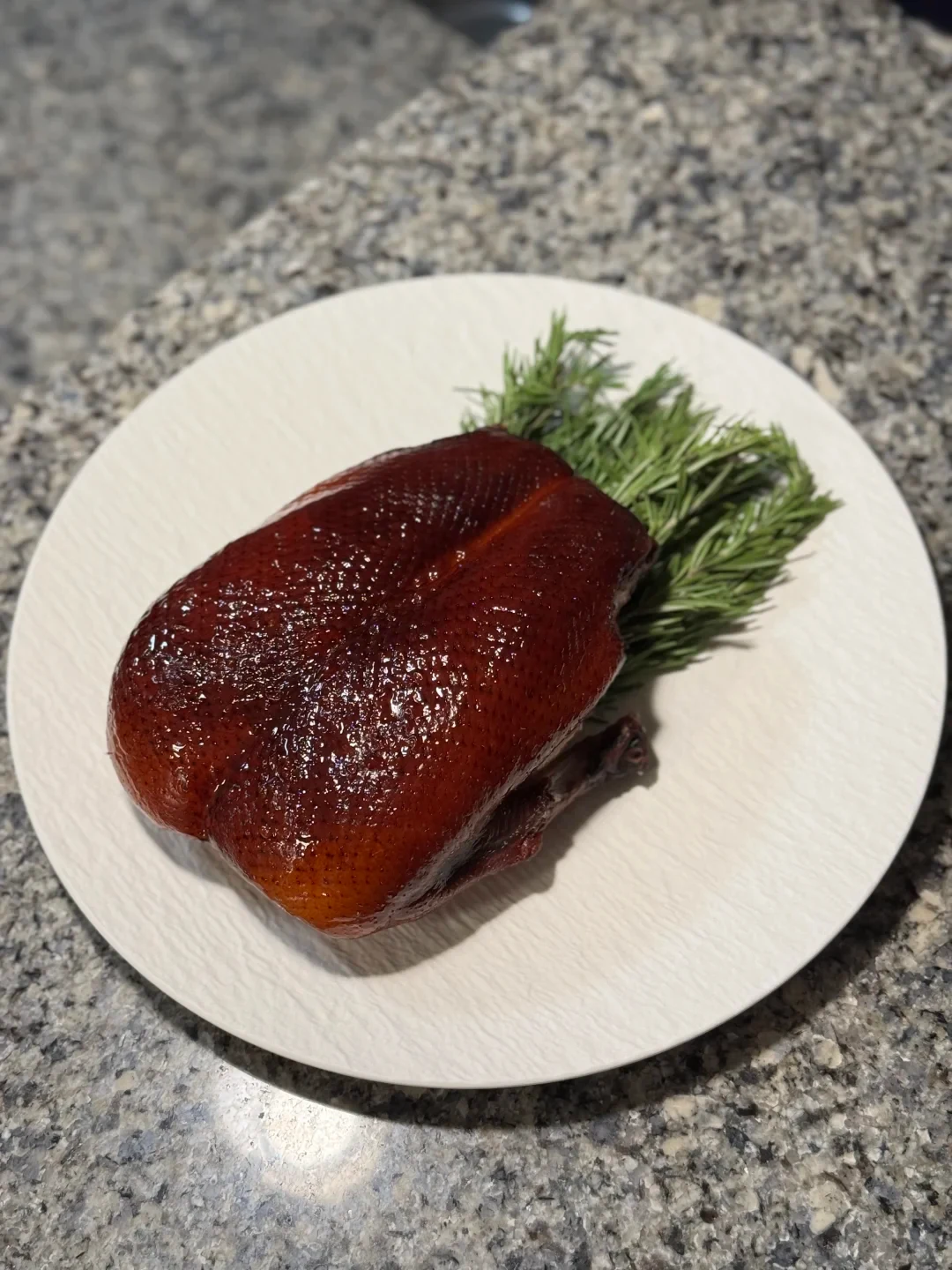 A glossy, dark reddish-brown roasted duck sits on a white plate with green herb sprigs behind it, on a speckled stone countertop.