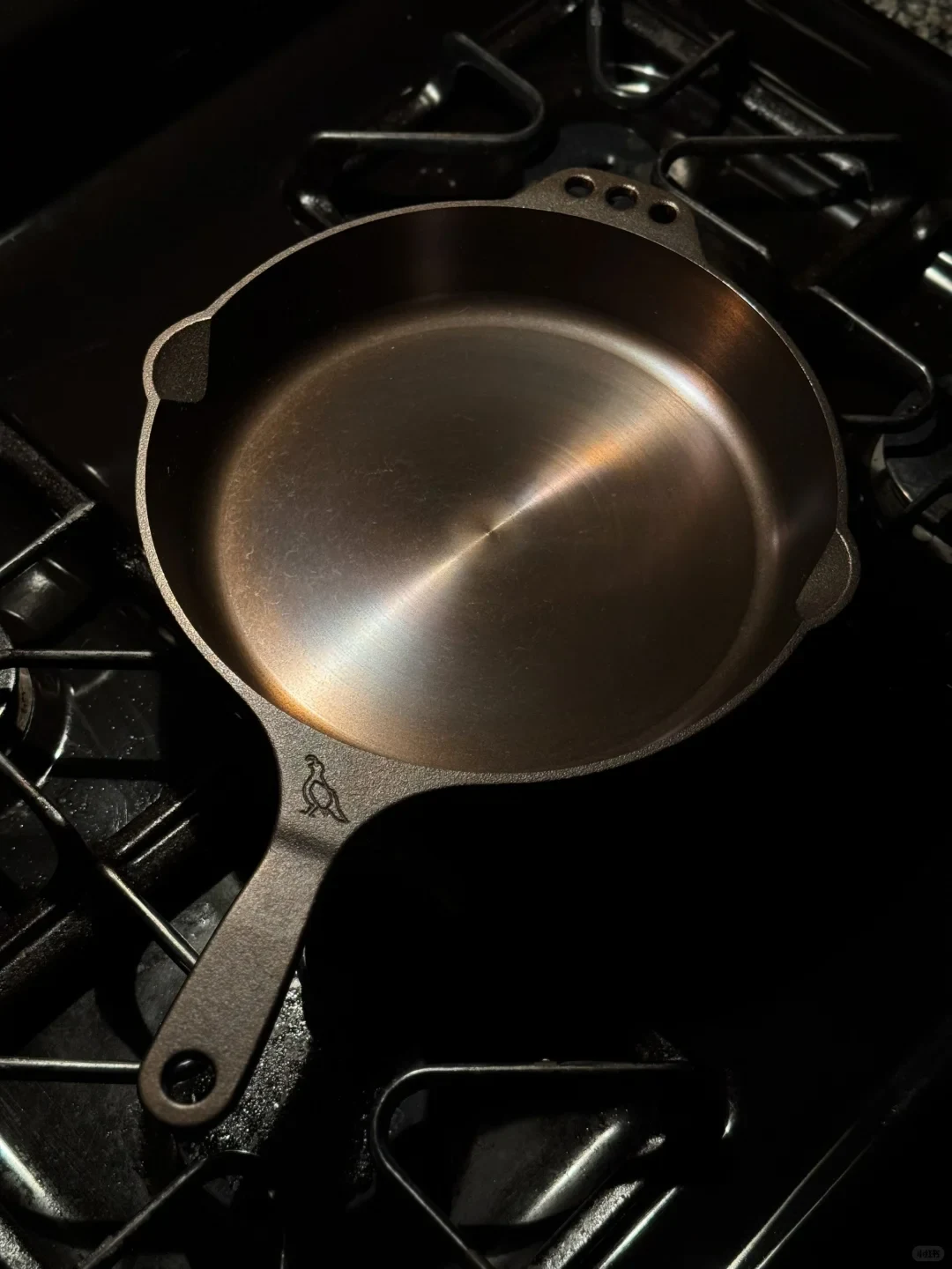 A dark metal frying pan with a long handle sits on the grate of a gas stovetop; the pan is empty.