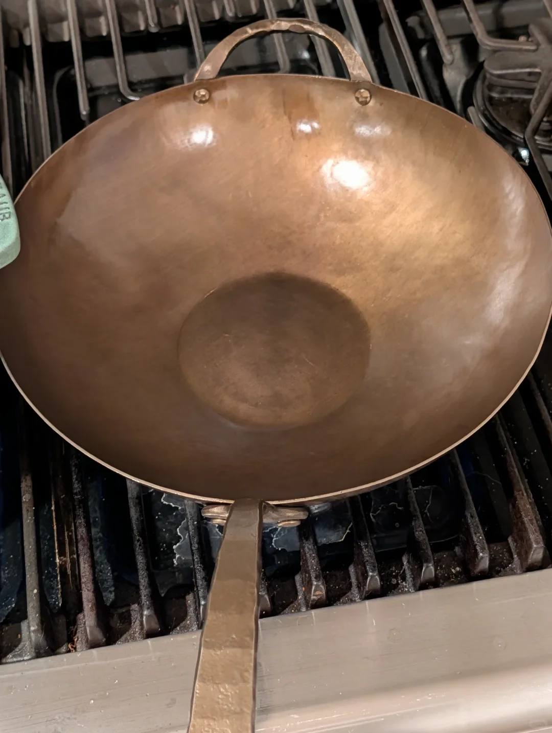 An empty, bronze-colored, hand-forged carbon-steel wok rests on stove grates, with a long flat handle toward the camera and a loop helper handle at the back.