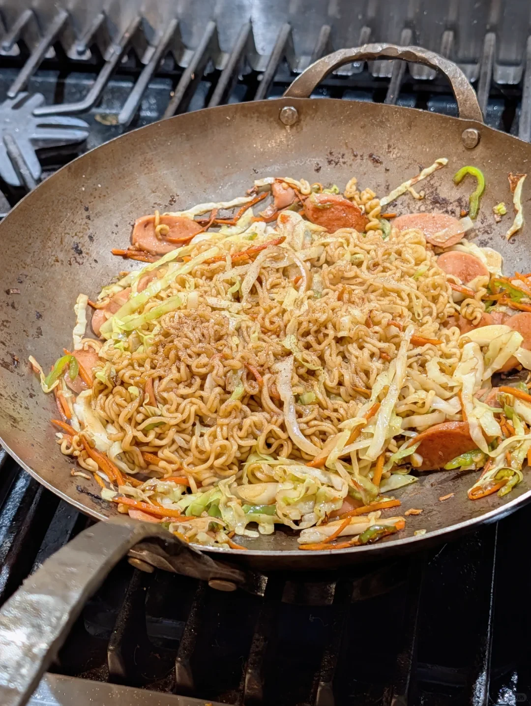 A carbon-steel wok on gas-stove grates holds stir-fried noodles with cabbage, carrot, green pepper and sliced sausage.