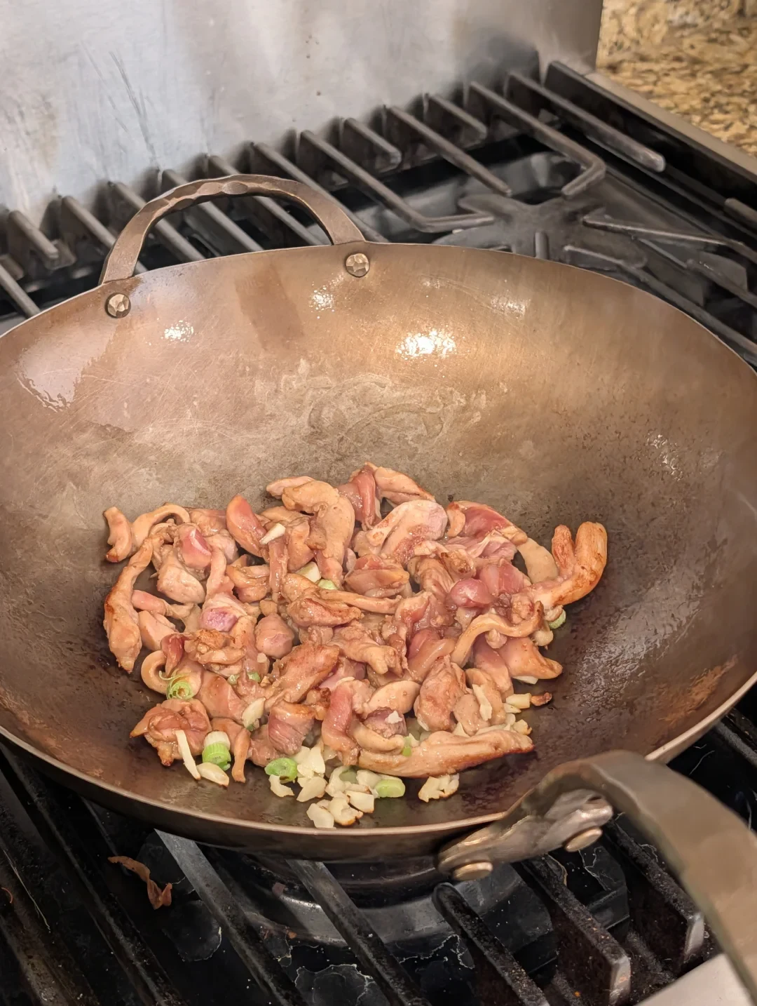 A hammered carbon-steel wok on gas-stove grates holds sliced meat with chopped scallions and pale aromatics.