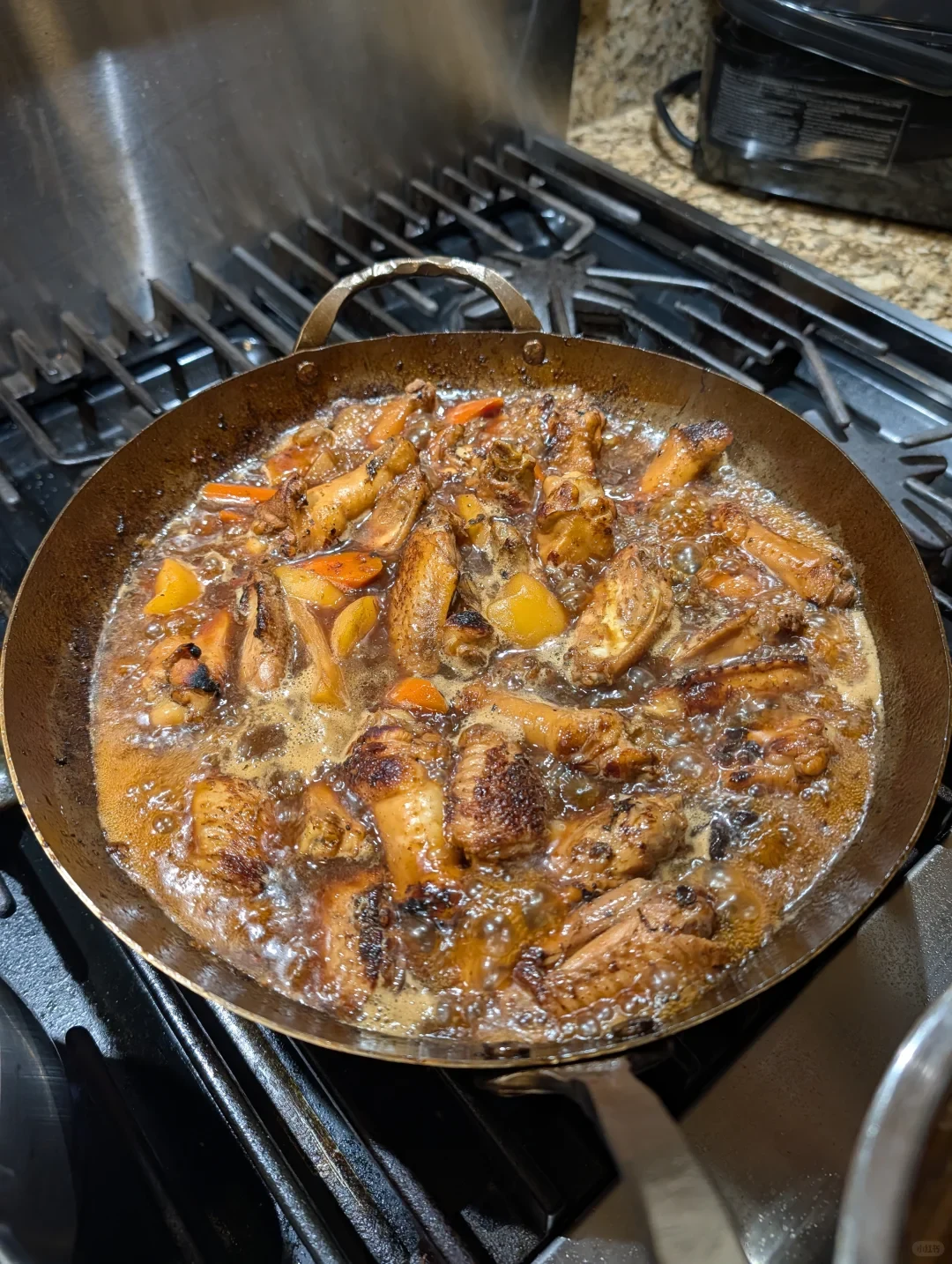 A pan of browned chicken pieces and orange vegetables simmers in bubbling sauce on a gas stovetop.