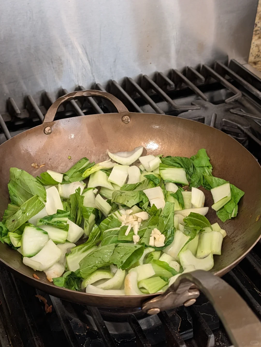 A metal wok on a gas stove contains chopped leafy greens and pale stems, with minced garlic scattered on top.
